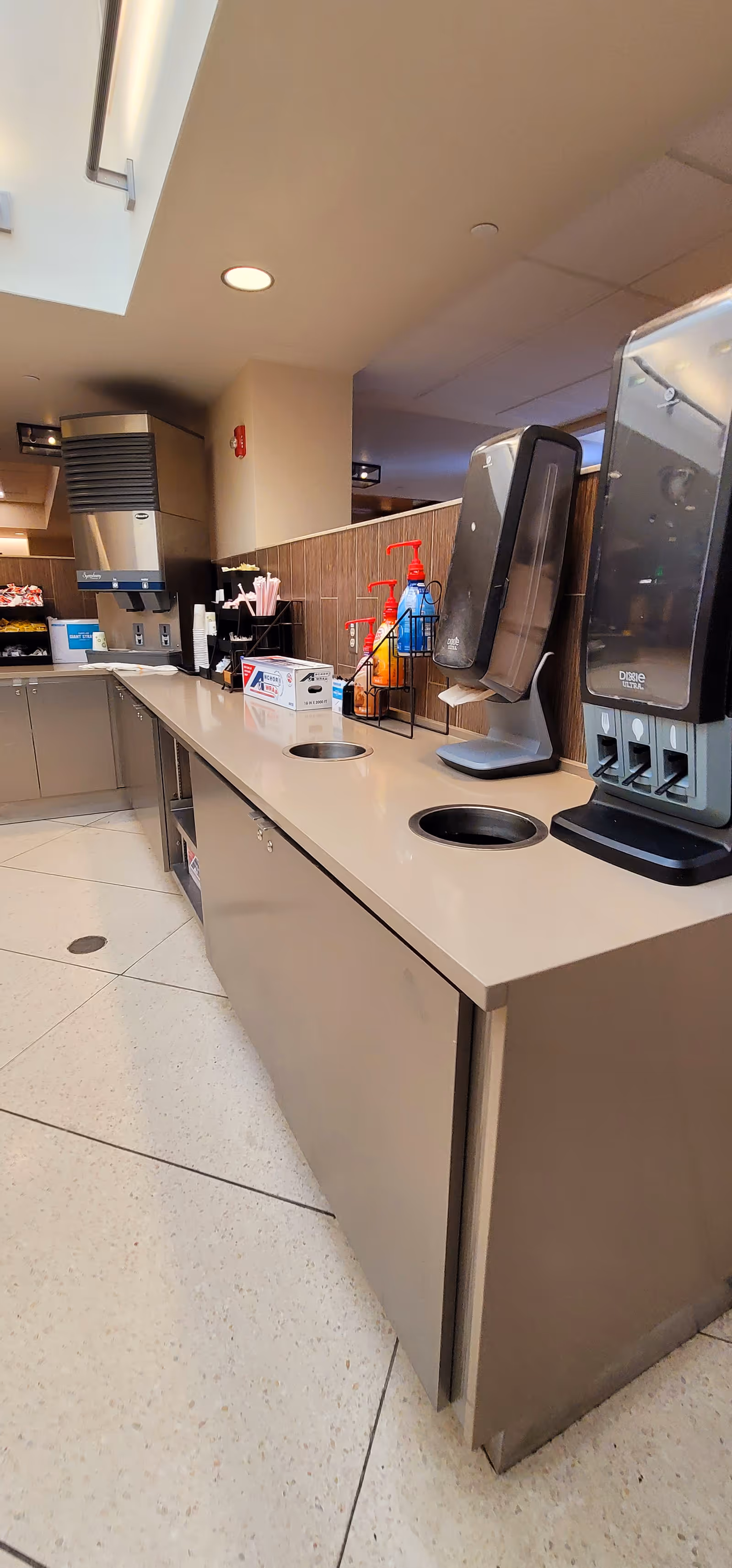 Long stainless service counter with beverage dispensers and cleaning supplies in an interior dining area.