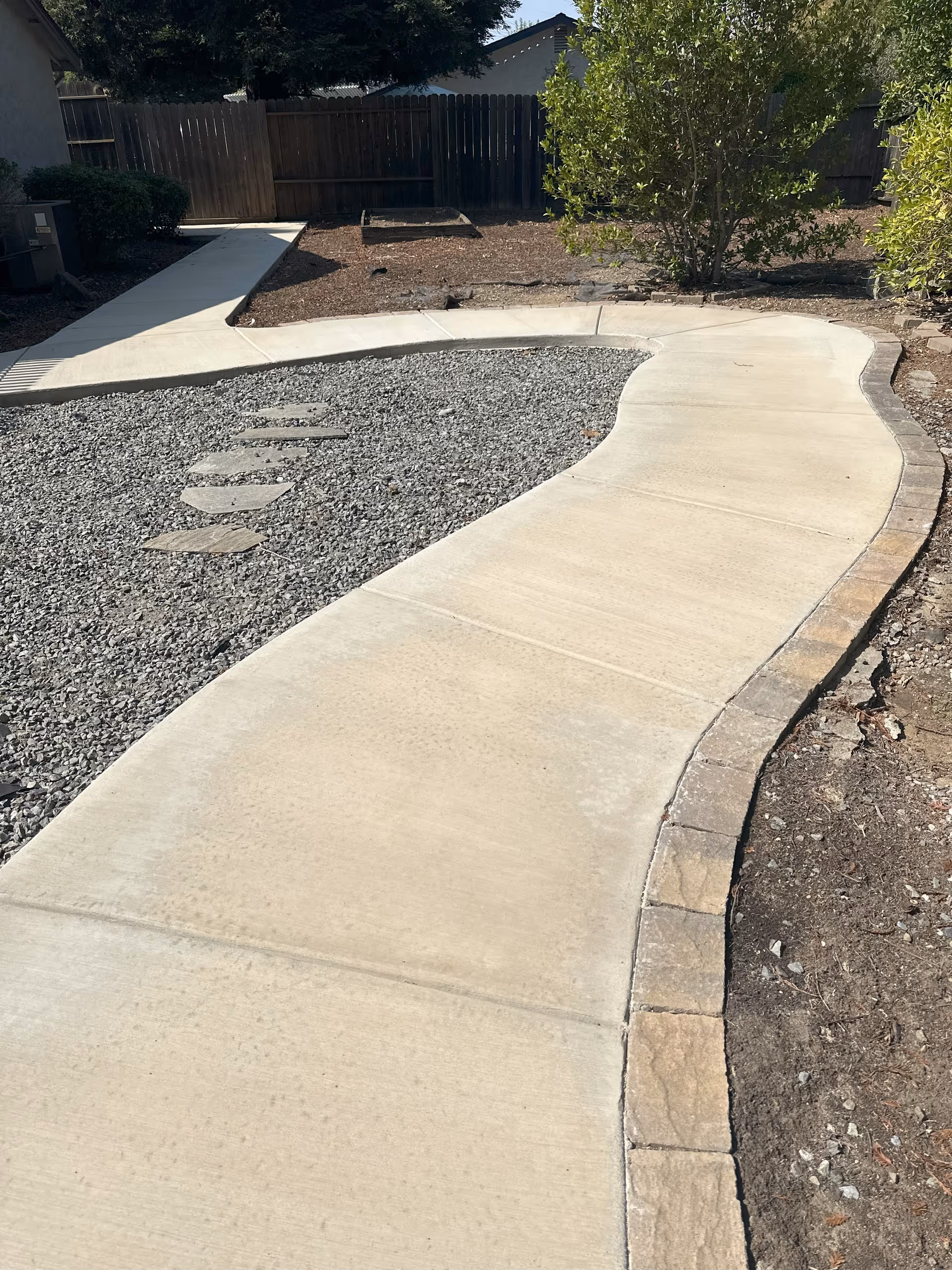 Curved concrete walkway through a landscaped yard with gravel and trees.