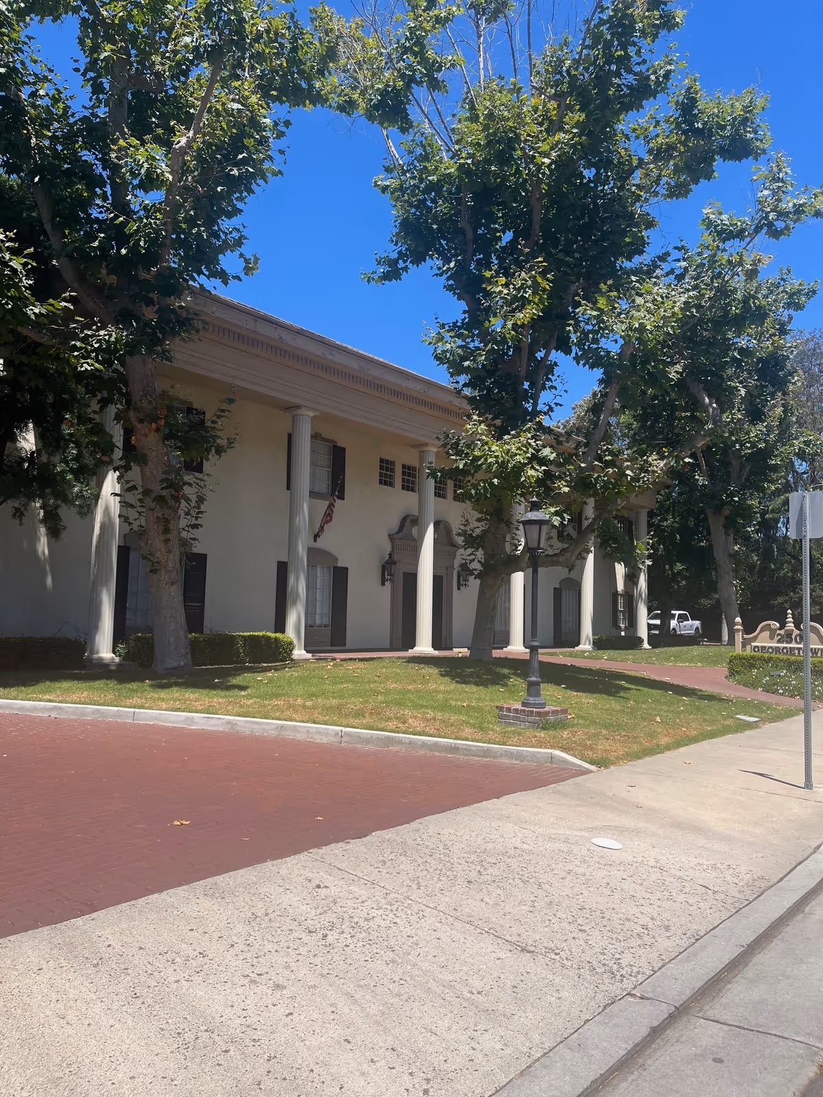 The front exterior of a building with columns, trees, and a sidewalk on a sunny day.