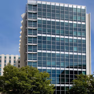 A modern glass-front building towers over trees against a blue sky.