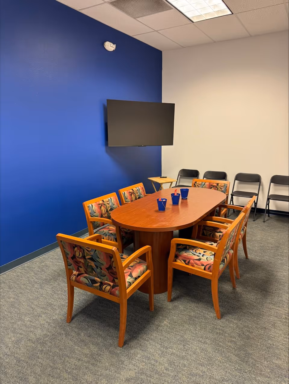 Small conference room with a wooden table, colorful chairs, and a wall-mounted TV.