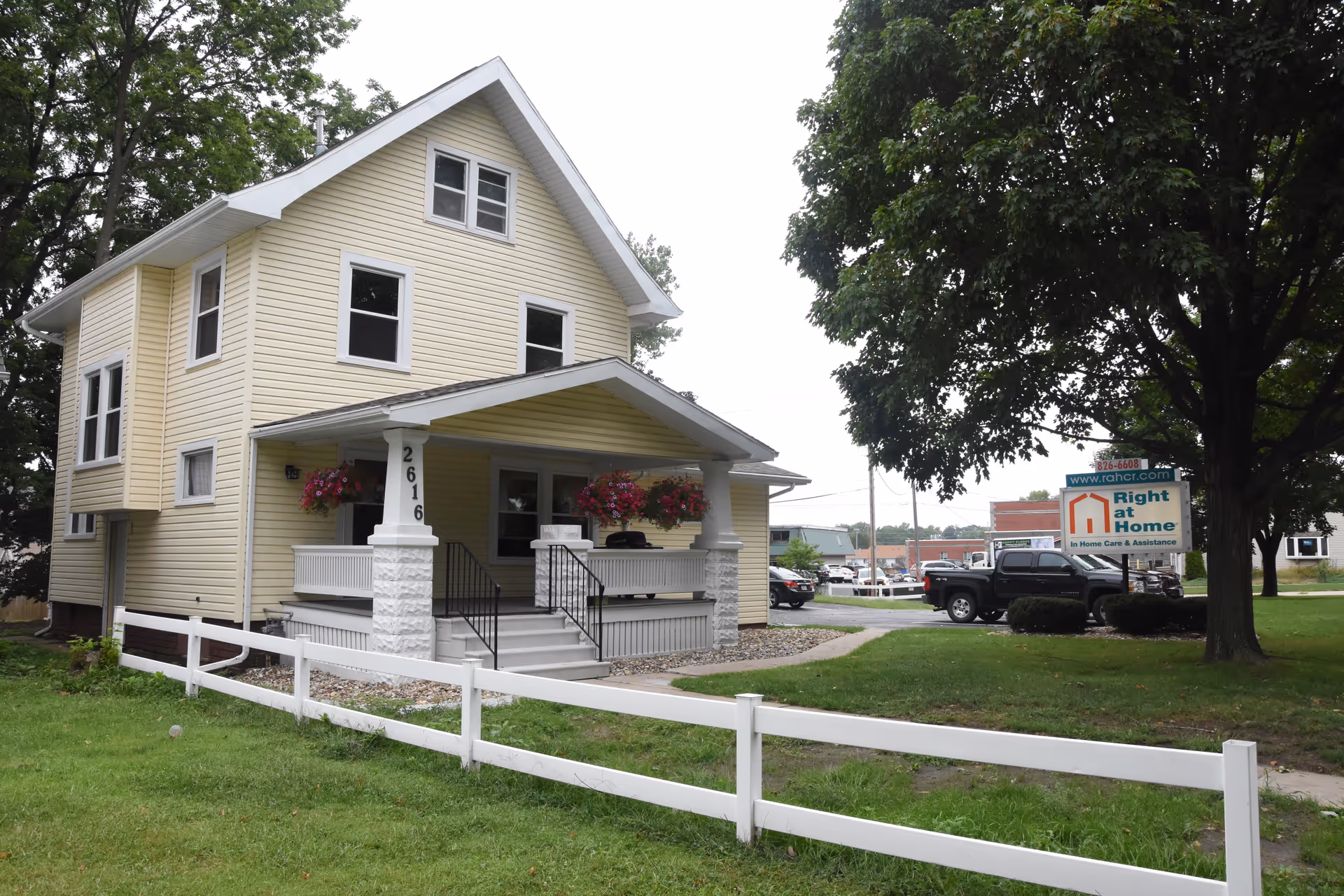 Yellow two-story residence with a front porch, flower baskets, and Right at Home sign.