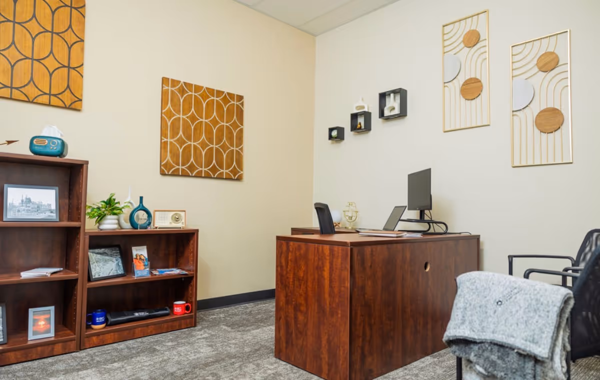 A wooden desk with computer and chairs, flanked by shelves and wall art.