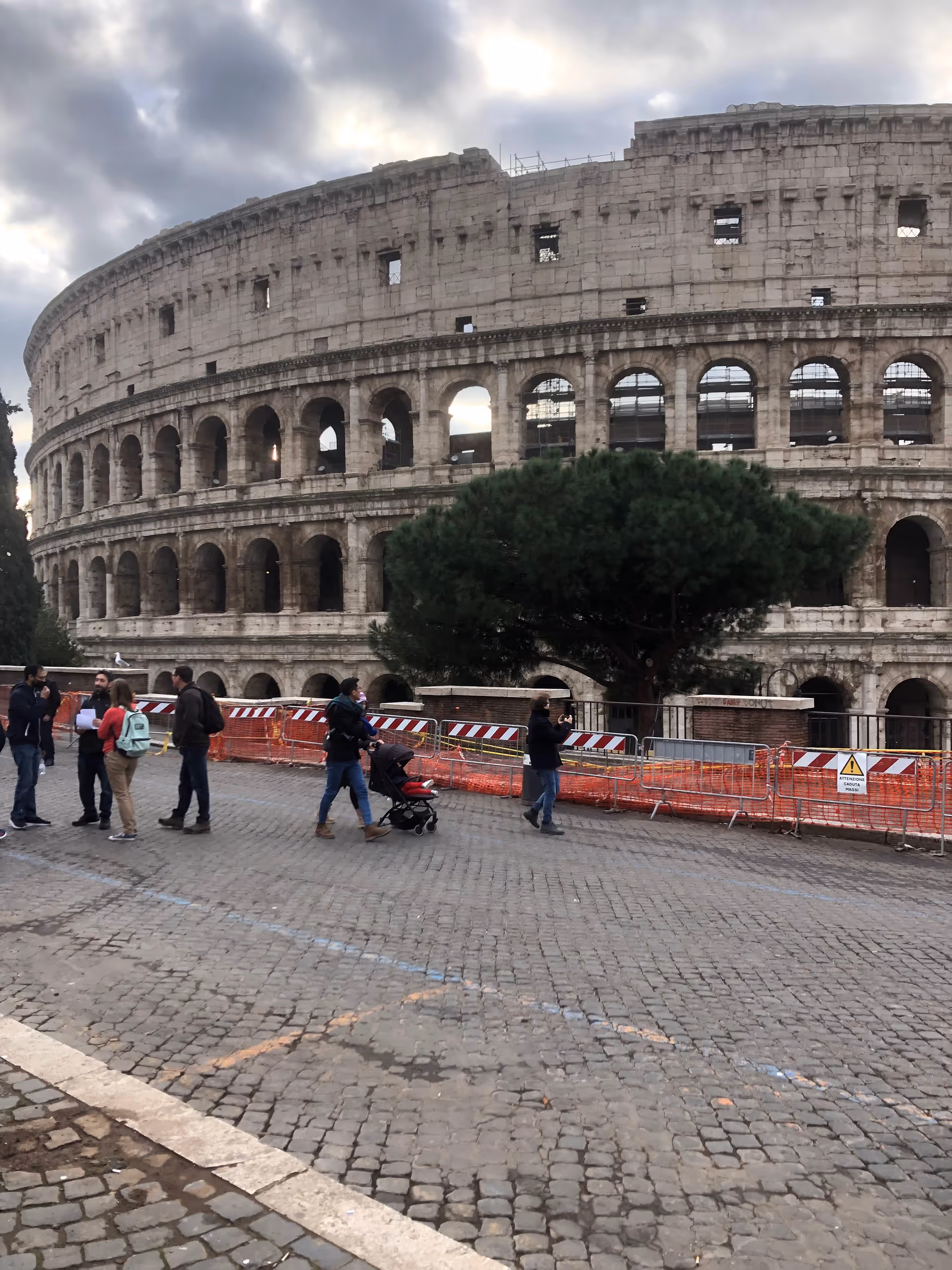 Tourists walk near the Colosseum with construction barriers on a cobblestone plaza.