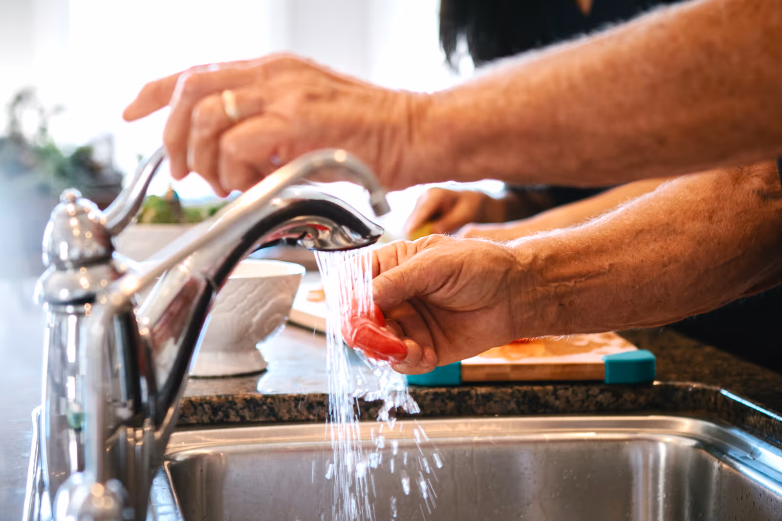 Close-up of hands washing a red vegetable under a kitchen faucet.