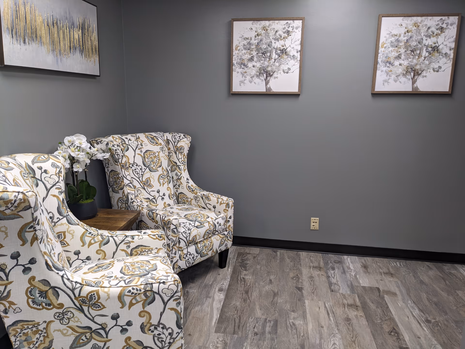 Two patterned armchairs with a wooden table and orchid, gray walls, framed art.