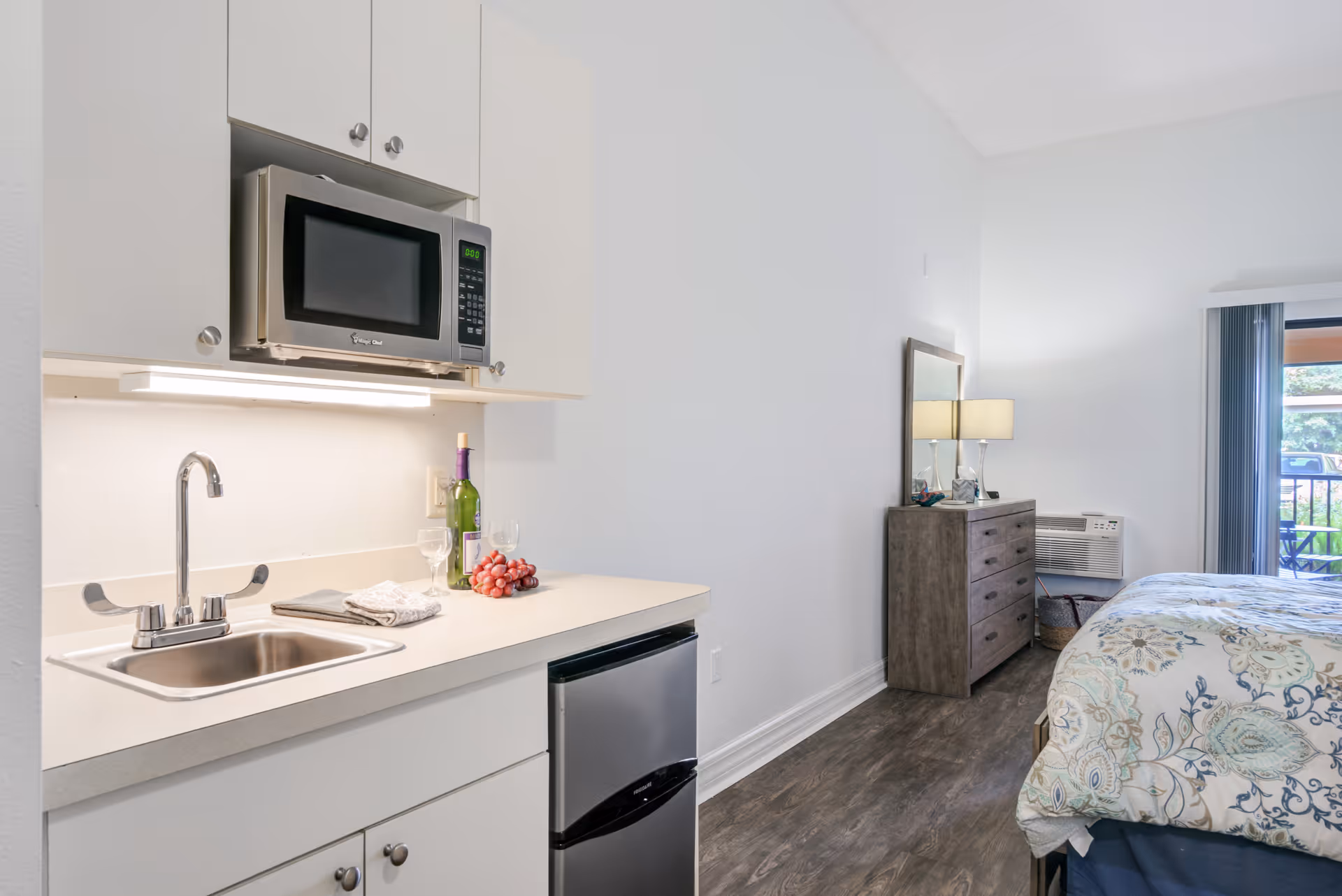 Compact kitchenette beside a bedroom with a dresser, bed, and balcony door.