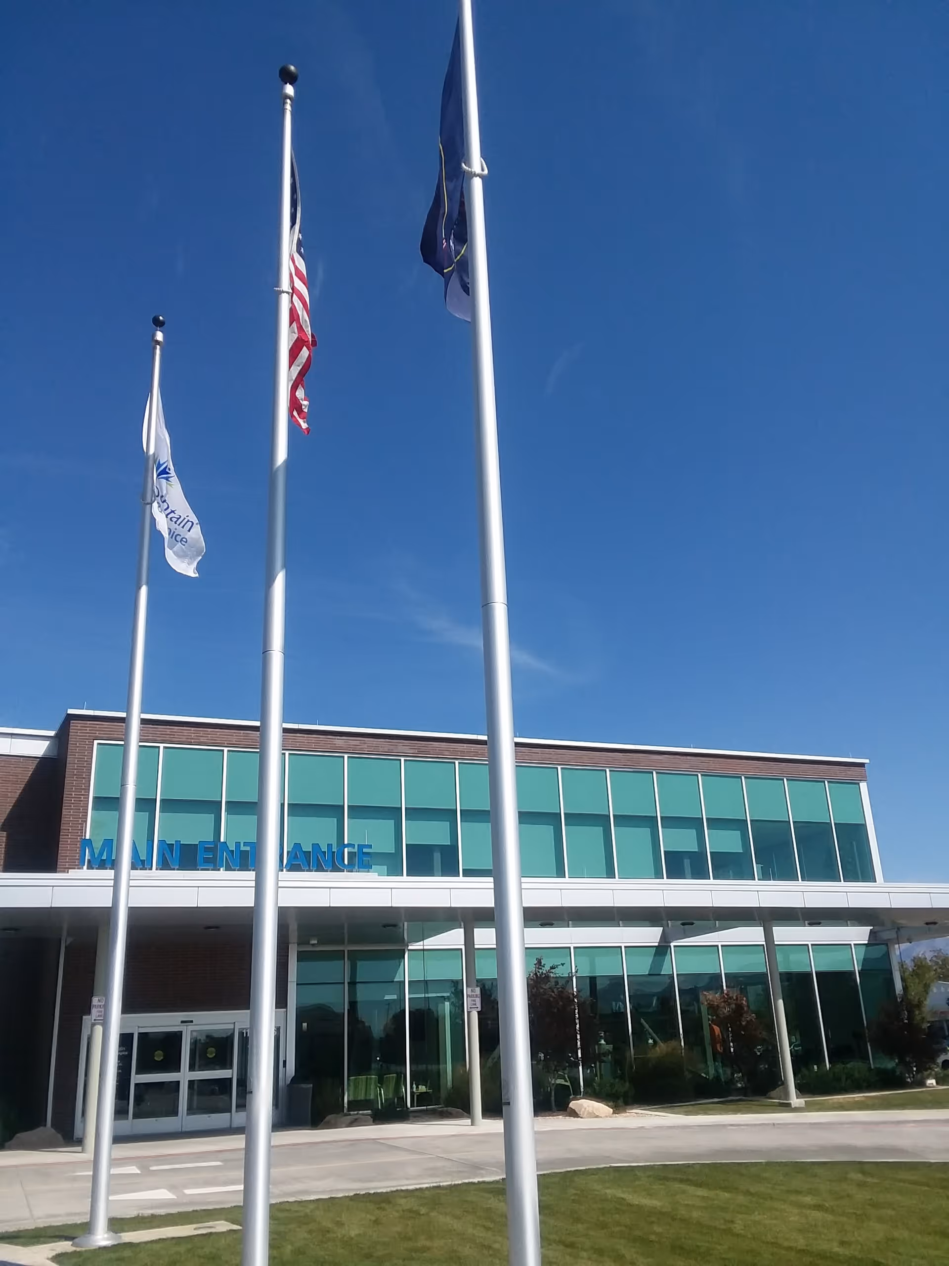 Modern glass-front facility with flagpoles, an accessible main entrance, and clear blue sky.