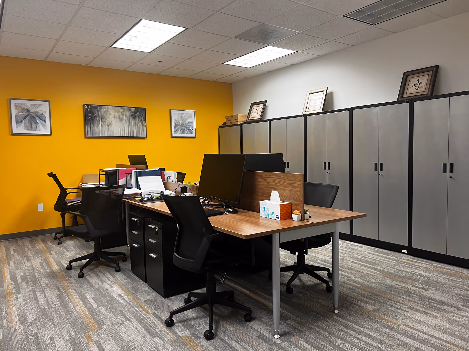 Office workspace with desks, chairs, computers, and gray cabinets along an orange accent wall.