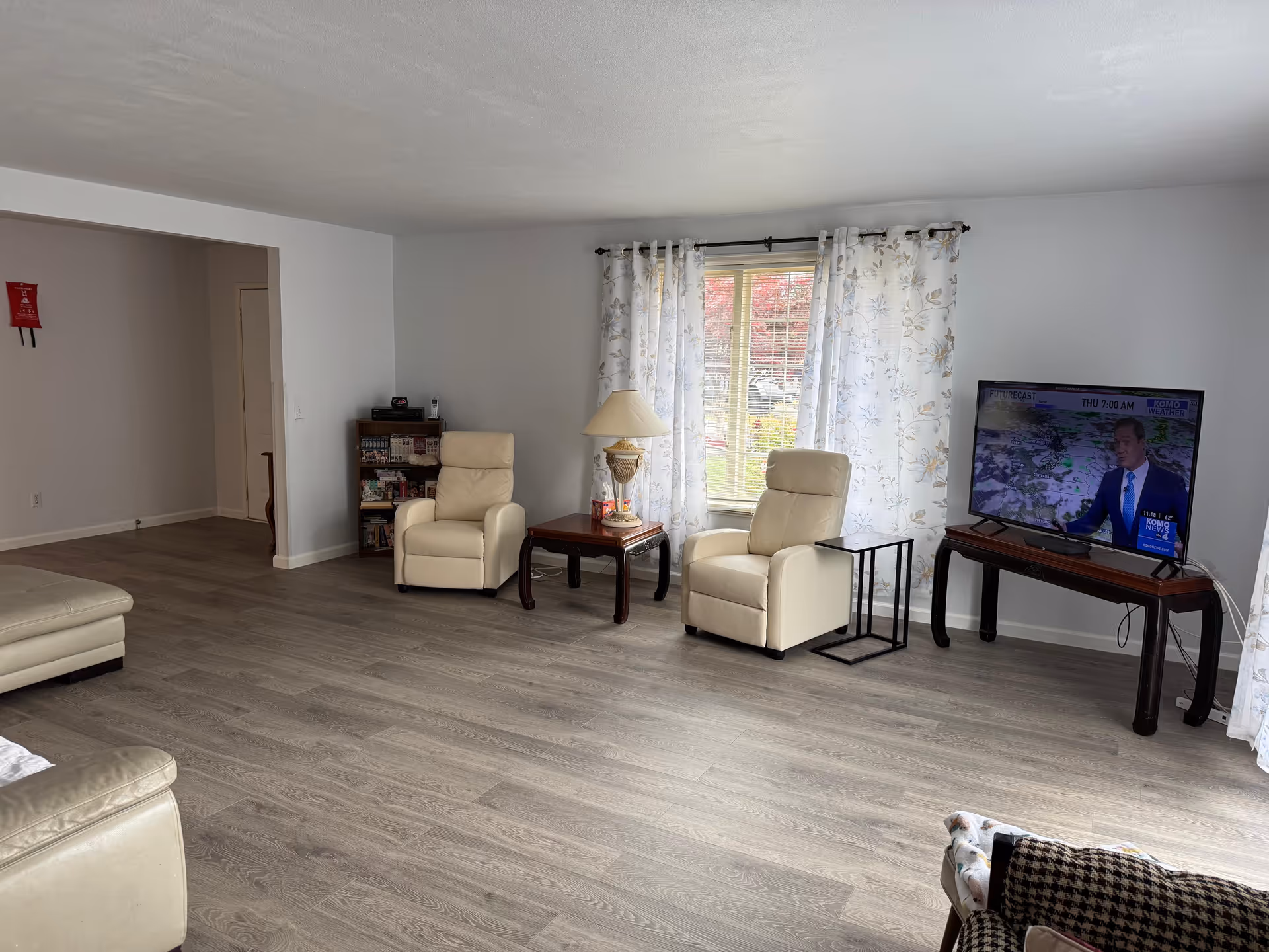 Spacious living room with beige recliners, a TV, lamp, bookshelf, and large window with curtains.