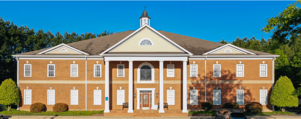 Two-story brick building with white columns, centered entrance, and surrounding green lawn.