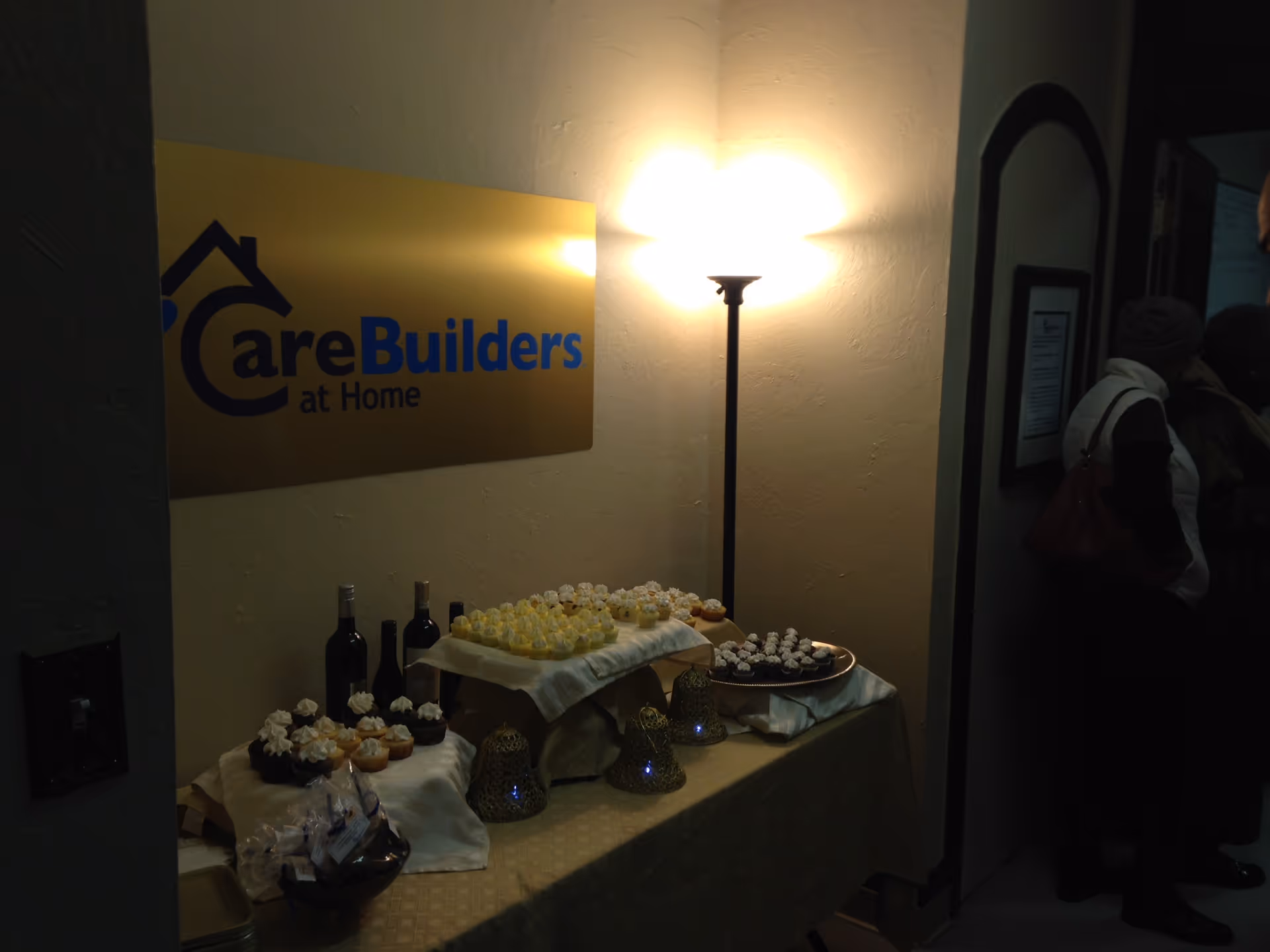 Dessert table with cupcakes and wine beside a CareBuilders at Home sign in a dim hallway.