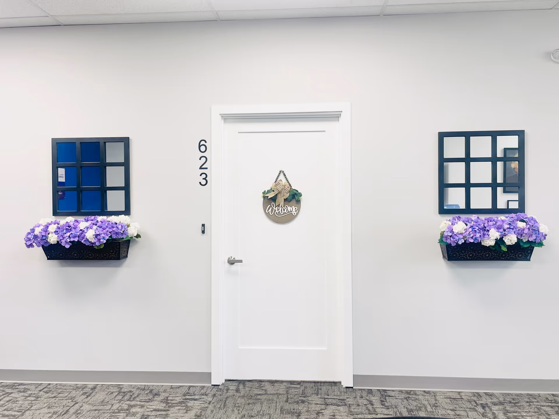 White interior hallway with central door, wreath, and purple flowers in window boxes.
