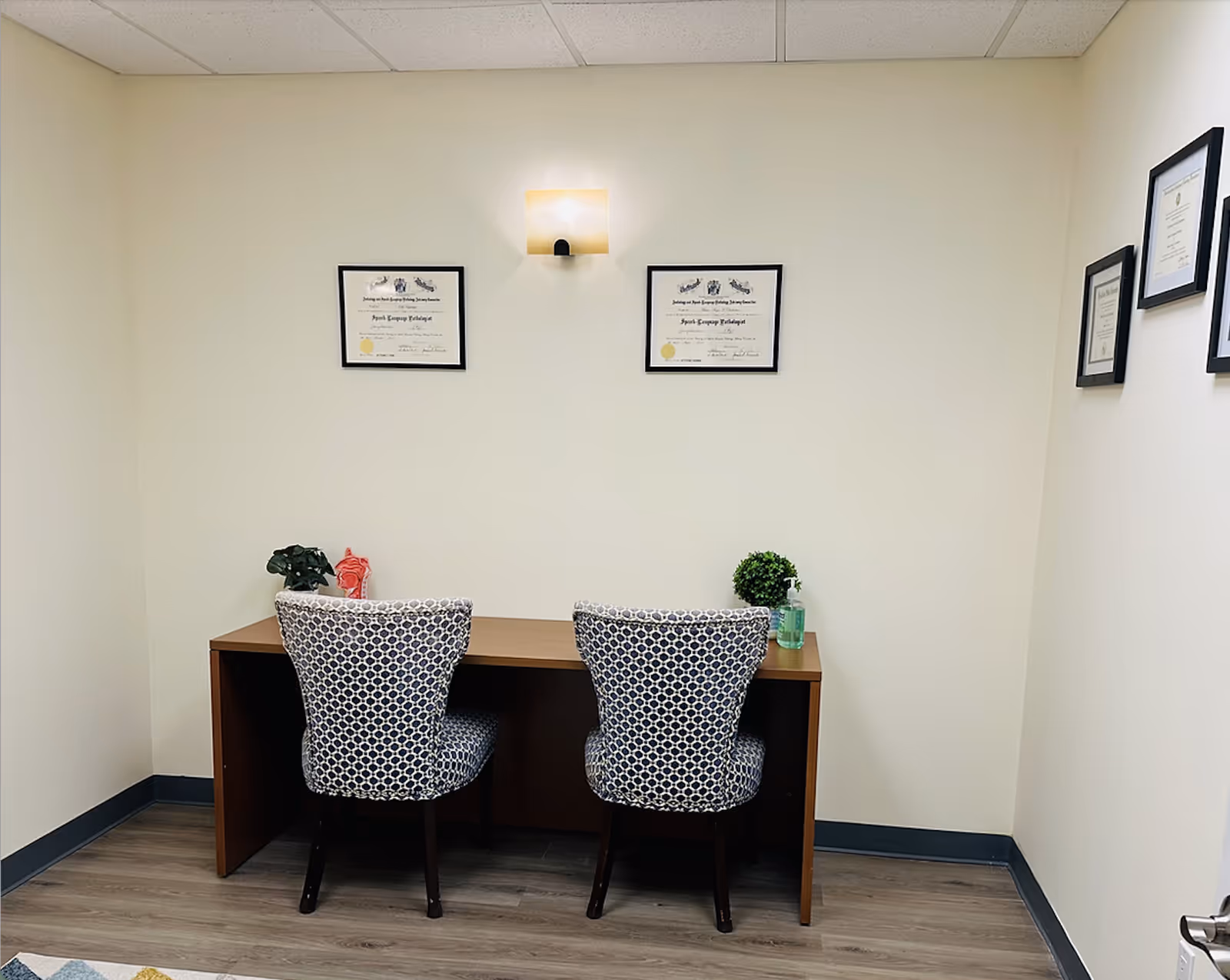 A quiet office space with a desk, two patterned chairs, and framed certificates.