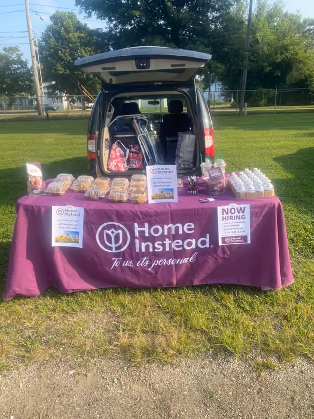 Open car trunk behind a purple Home Instead table with snacks outdoors.
