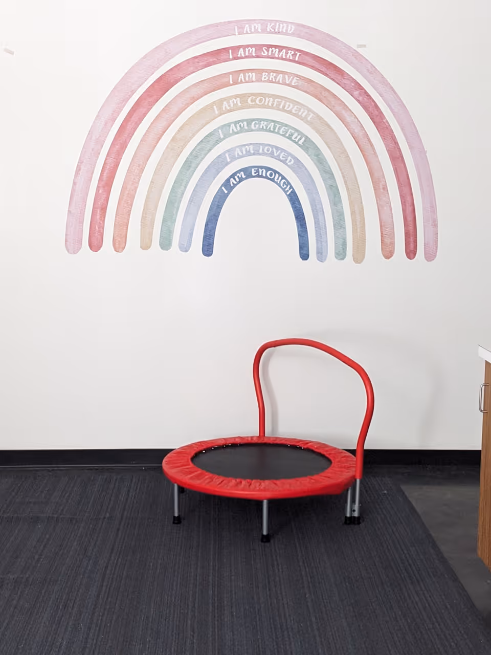 Small red exercise trampoline with a curved handle in front of a pastel rainbow mural.