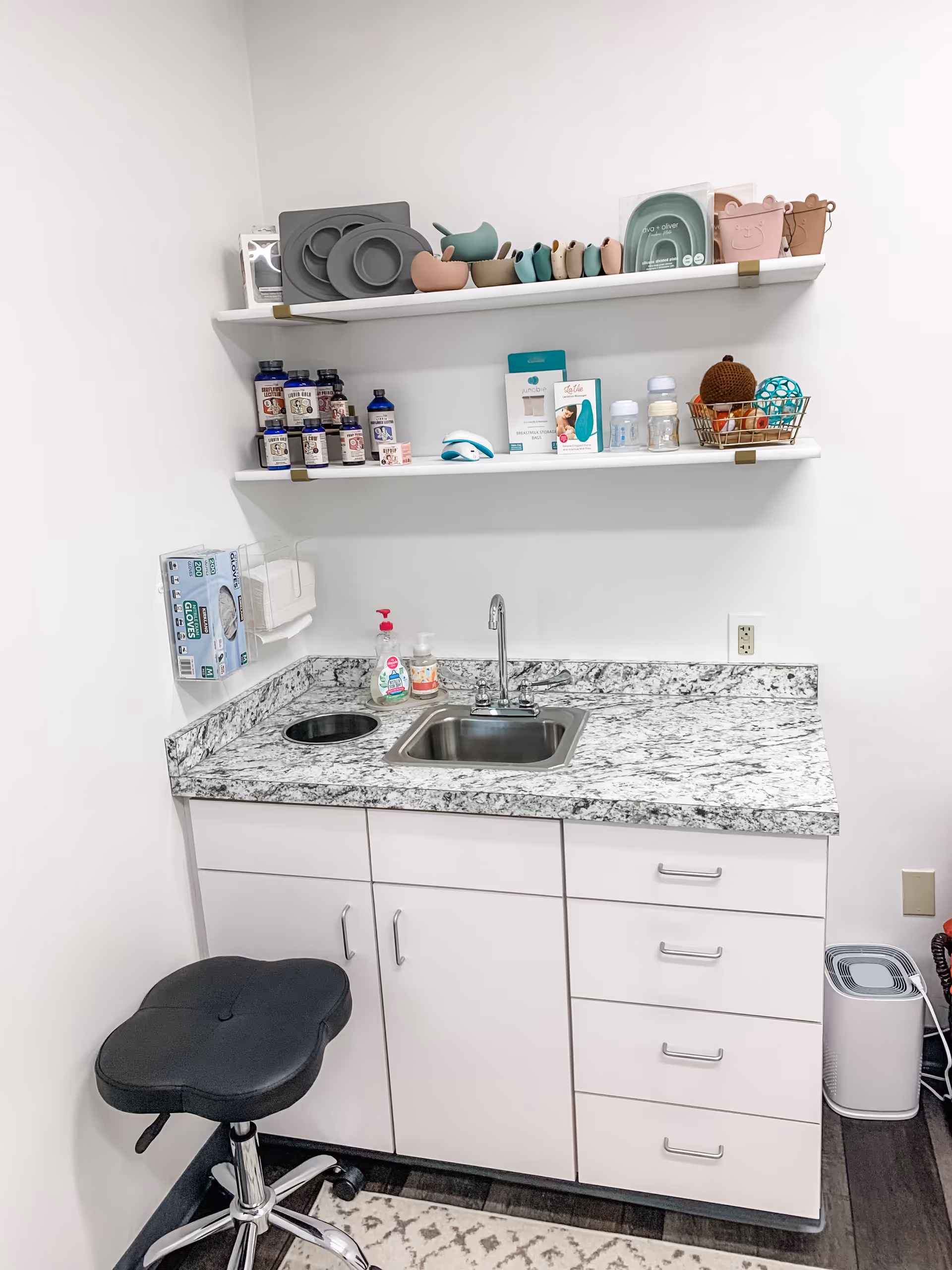 Small white kitchenette with granite countertop, sink, shelves holding bottles and baby items, and a stool.