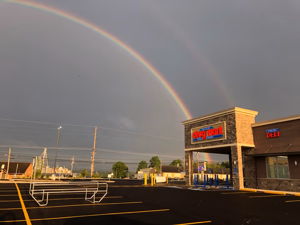 Double rainbow arches over Discount Drug Mart storefront with a parking lot.