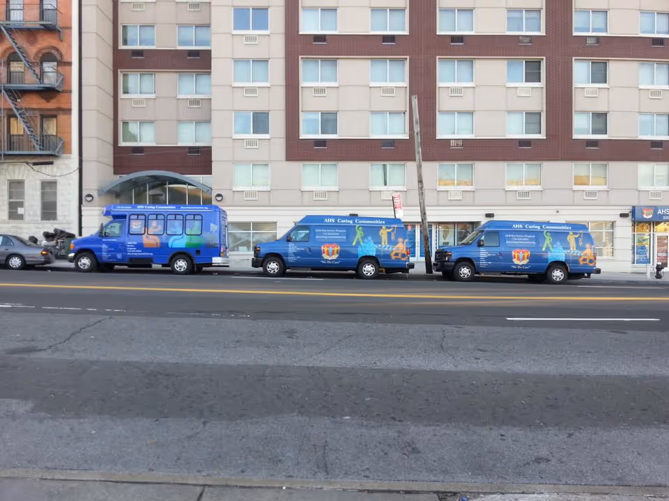 Blue vans with AHS Caring Communities parked along a city street in front of a building.