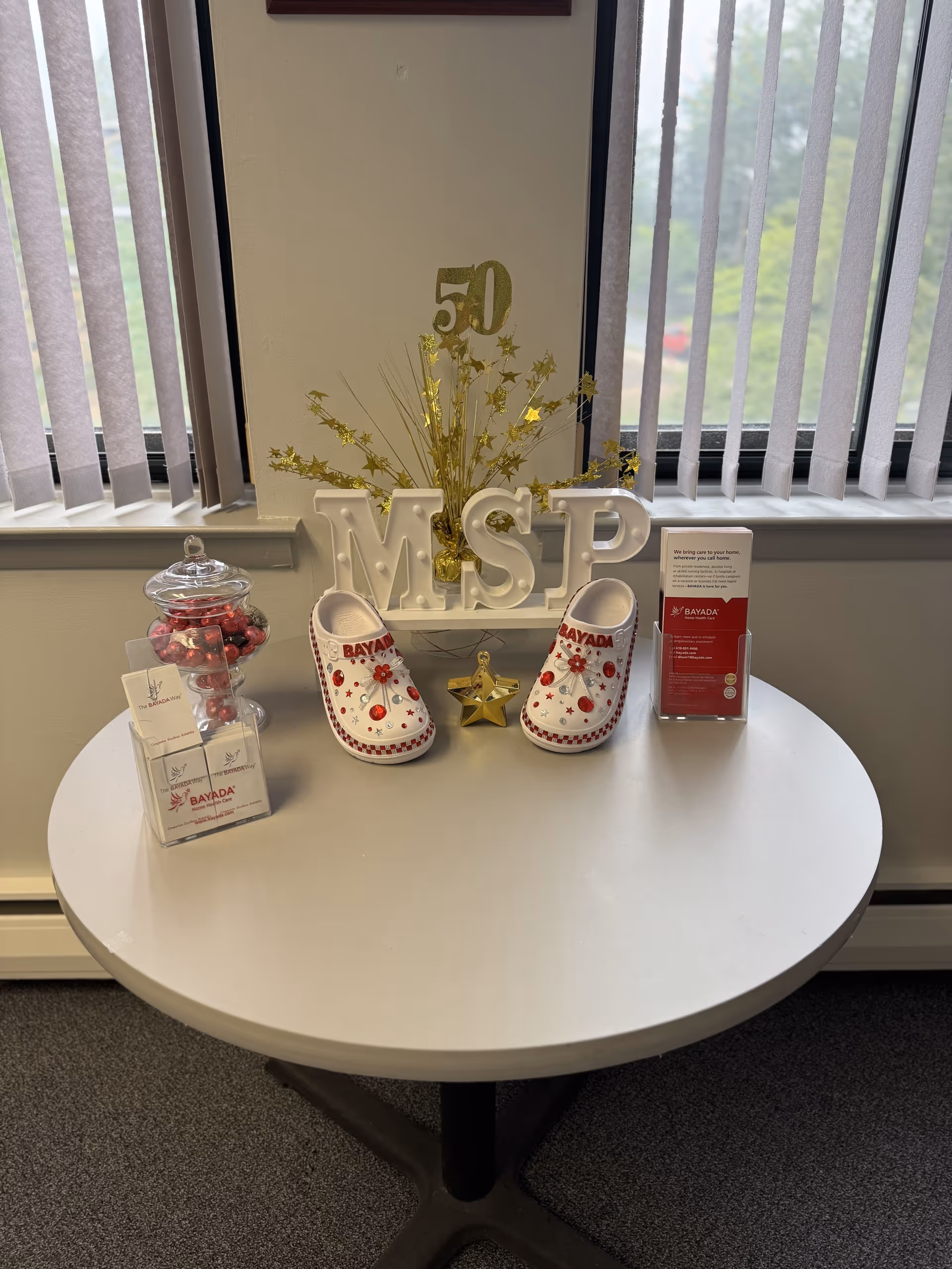Round table with BAYADA decor, decorative shoes, and a candy jar near window blinds.