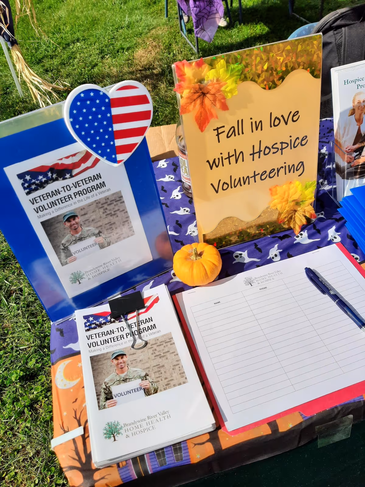 Outdoor table with hospice volunteer materials, sign-in sheet, and fall decorations.
