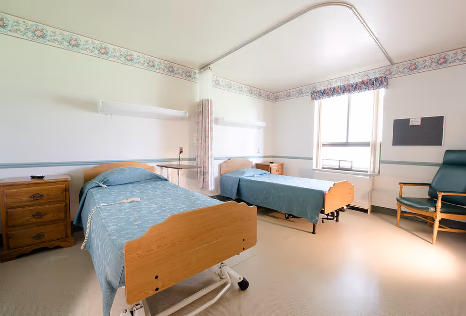 Two hospital beds in a pale patient room with blue bedspreads and a window.