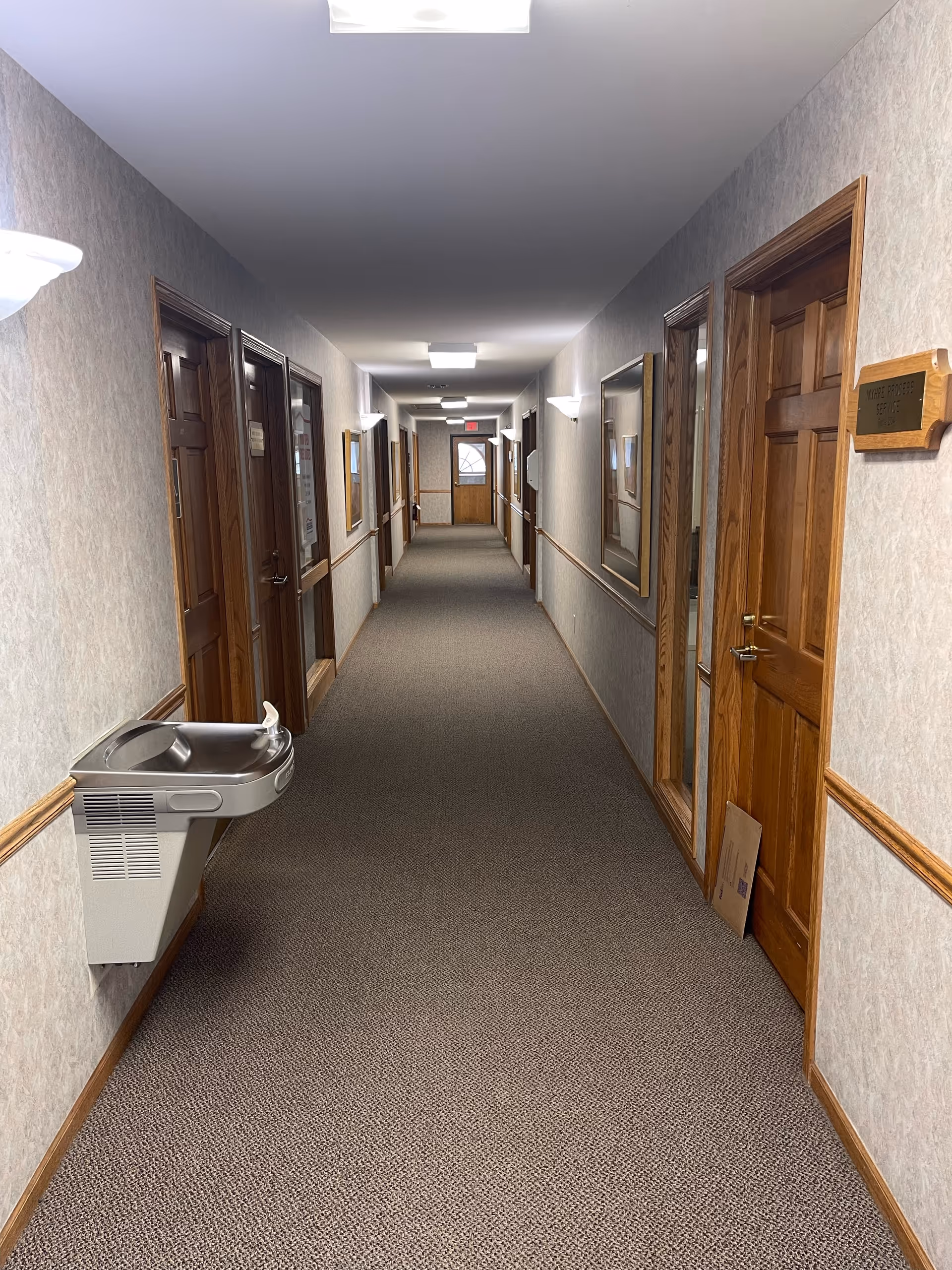 Long carpeted hallway in a senior living facility with wooden doors and wall-mounted fixtures.