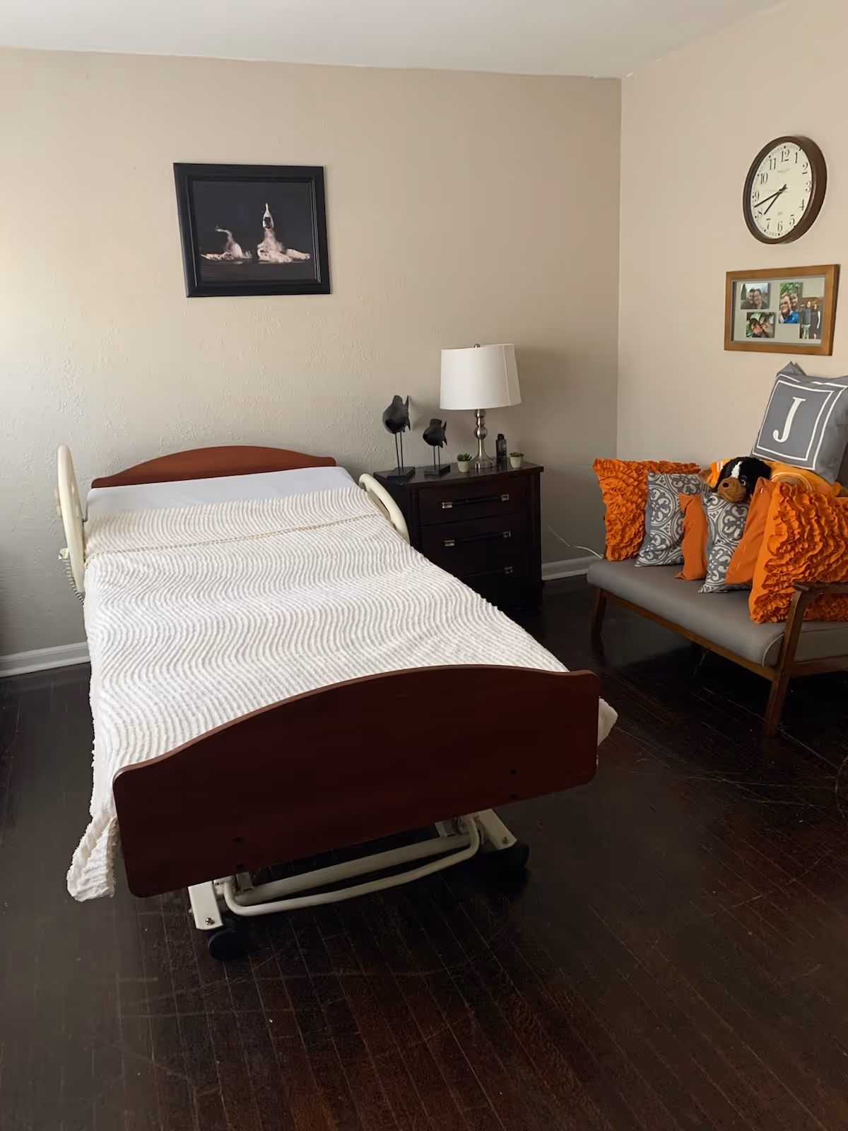 A furnished bedroom with an adjustable hospital-style bed, nightstand, lamp, and sofa.