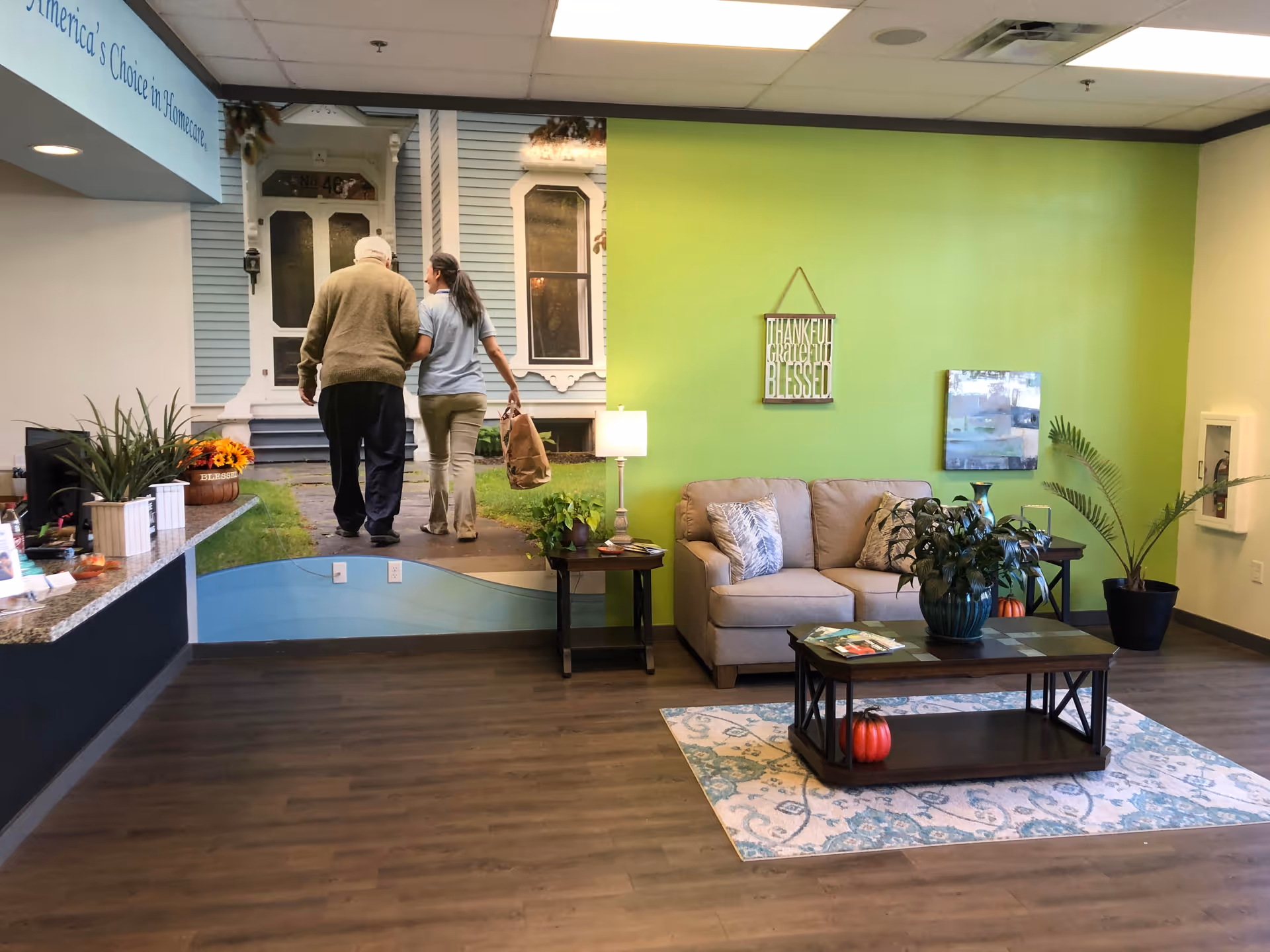 Indoor senior living lobby with sofa, coffee table, plants, and exterior house mural.