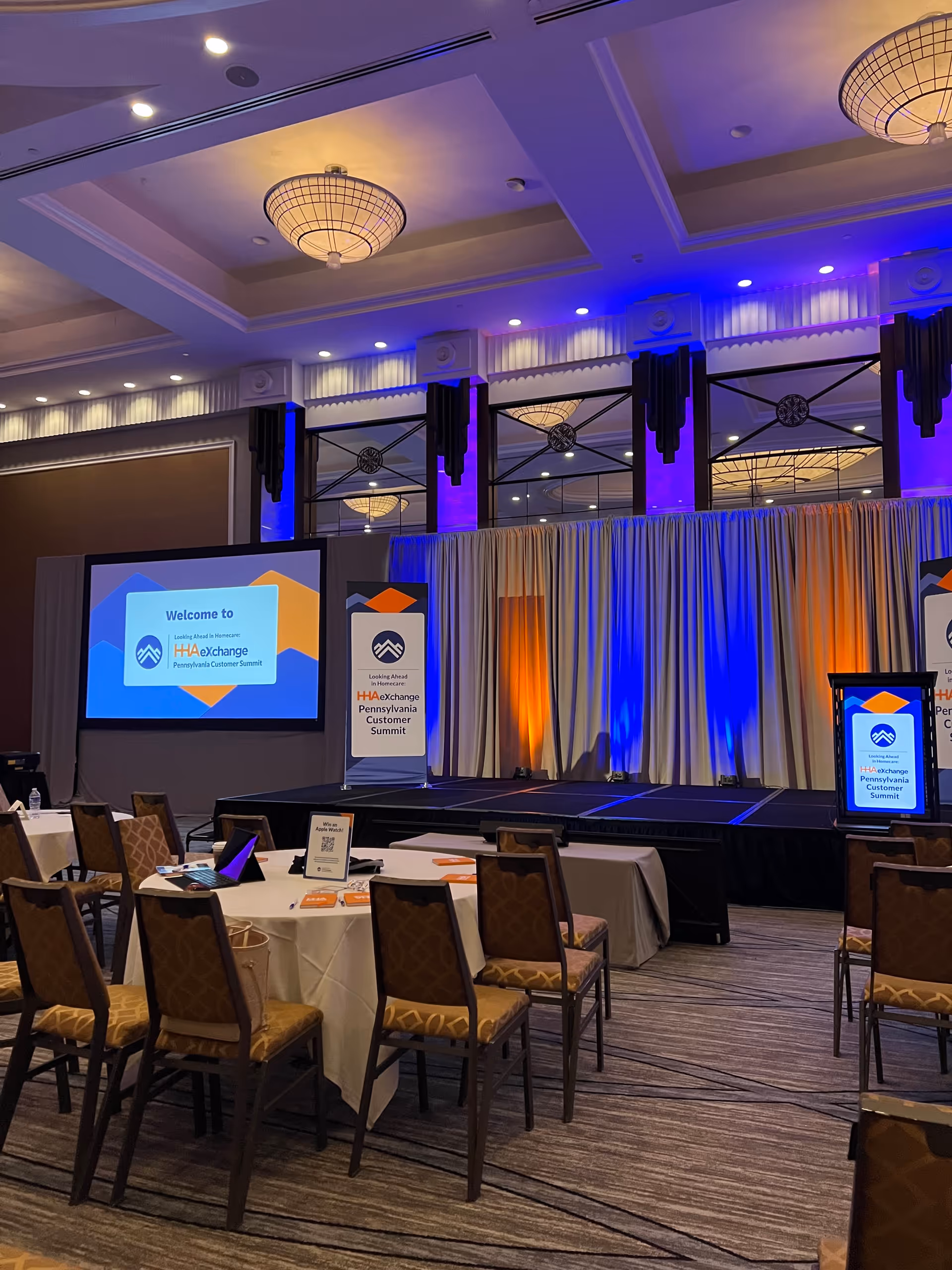 Indoors conference hall with stage, screens, banners, and arranged tables.