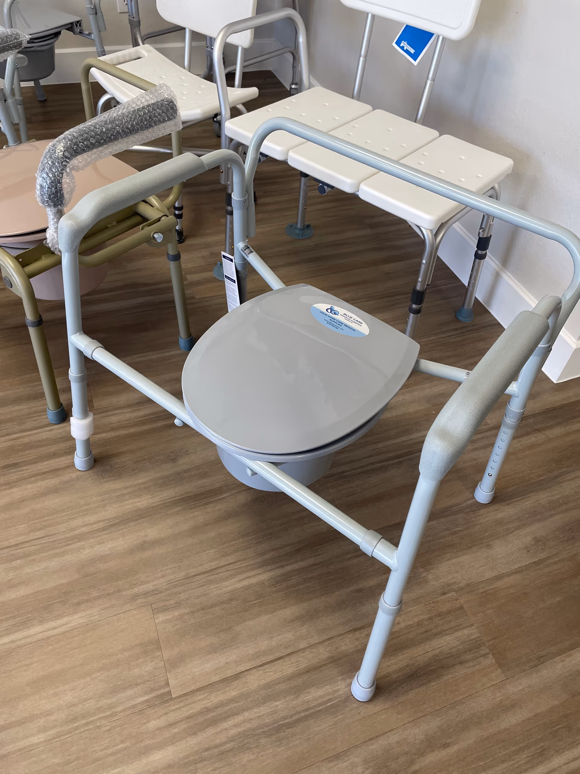 Assisted living commode chair and transfer chairs in a clean, wood-floored interior.