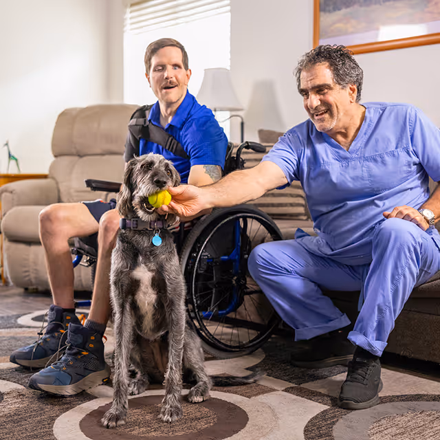 A caregiver in scrubs interacts with a resident and a dog in a cozy living room.