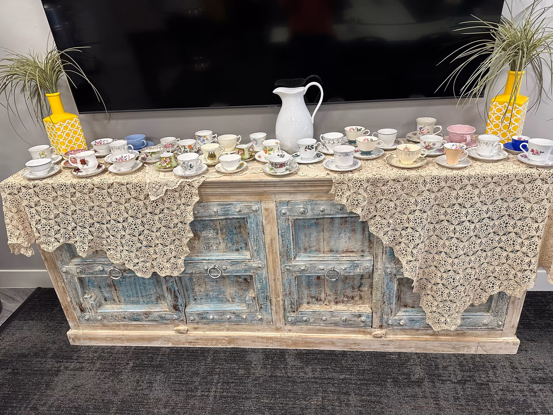 A vintage distressed cabinet displays teacups and saucers on lace runners with a white pitcher centerpiece.