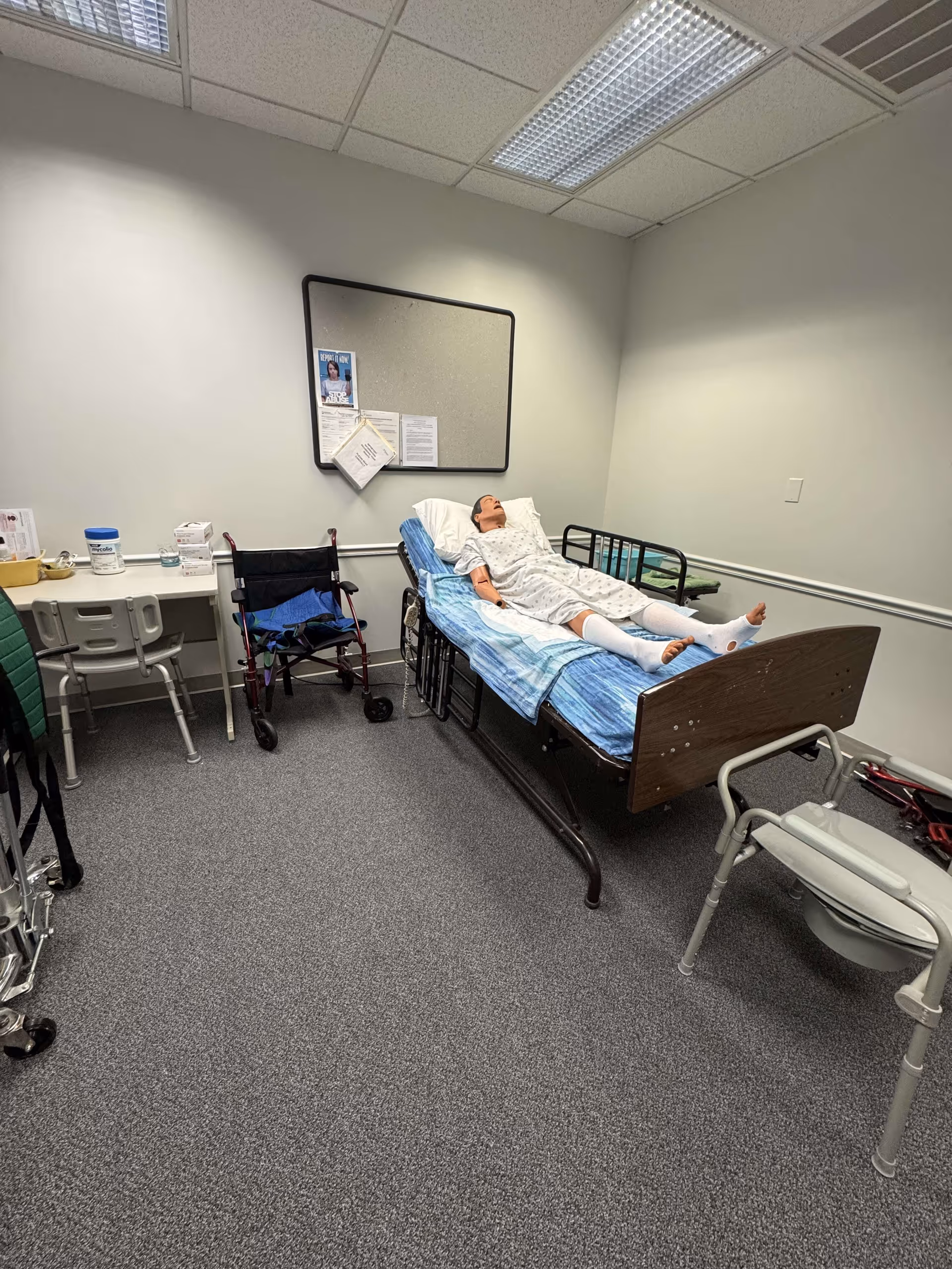 A hospital-style patient room with a person lying on a bed, medical cart and wheelchair nearby.