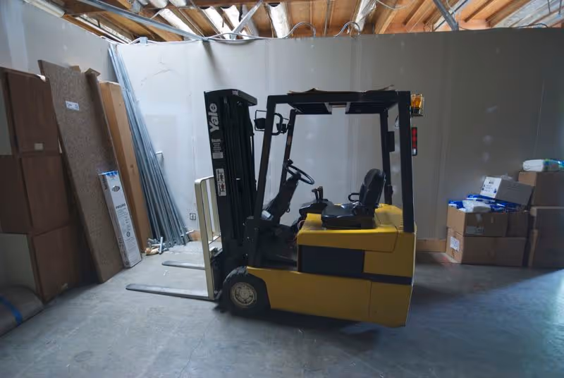 Yellow forklift in a cluttered storage room with boxes and metal panels.