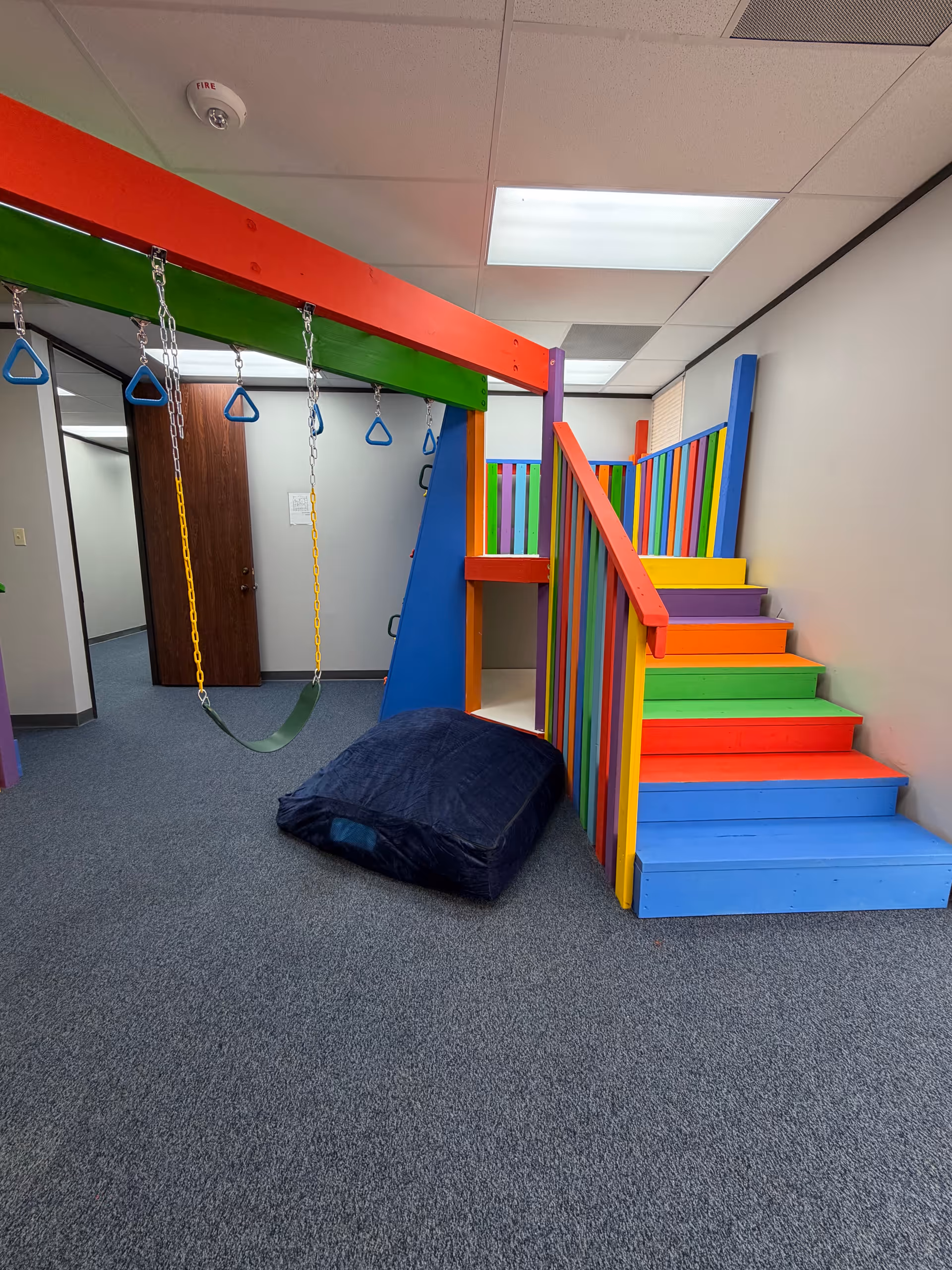 Colorful indoor play area with swings, padded stairs, and a vibrant climbing structure.
