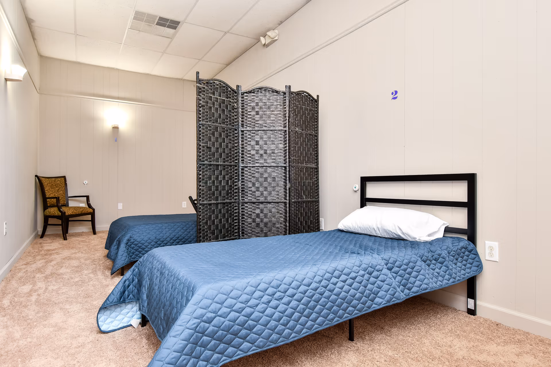 Two twin beds with blue quilts and a folding room divider in a neutral bedroom.