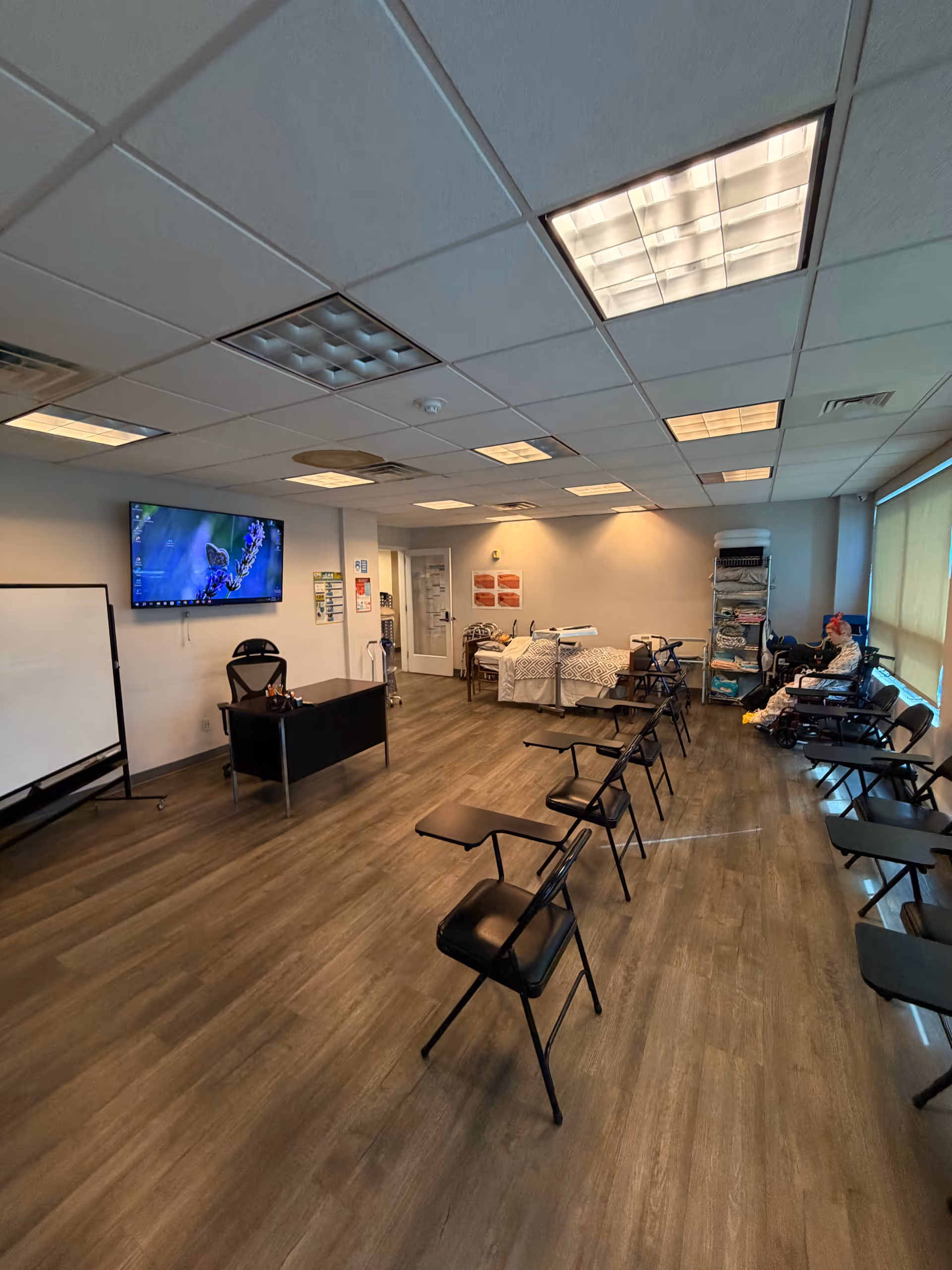 Spacious interior room with rows of black chairs, a desk, whiteboard, and wall-mounted TV.