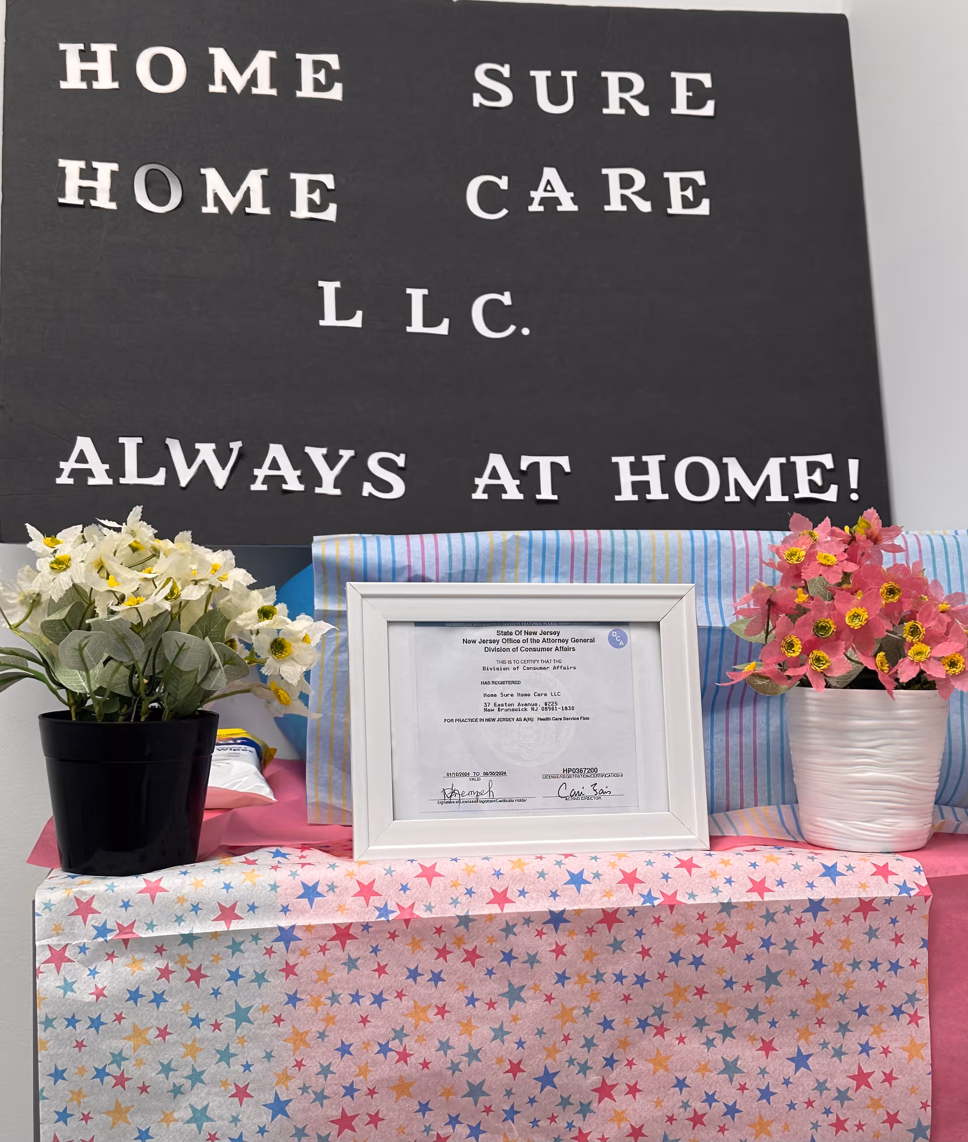 Interior display with a black sign HOME SURE CARE LLC ALWAYS AT HOME!, certificate, and flowers.