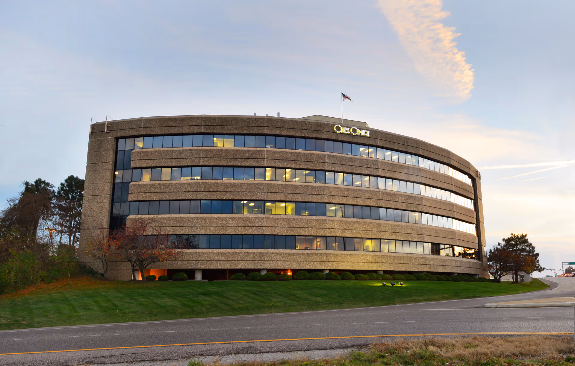 Curved brick building with large windows sits beside a road on a grassy lawn.