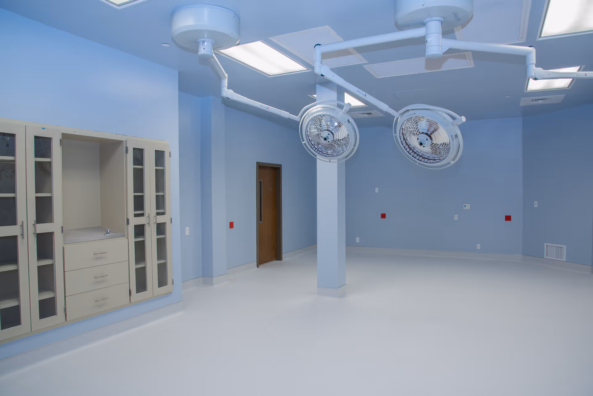 An empty hospital operating room with blue walls, ceiling-mounted lights, and large surgical lamps.