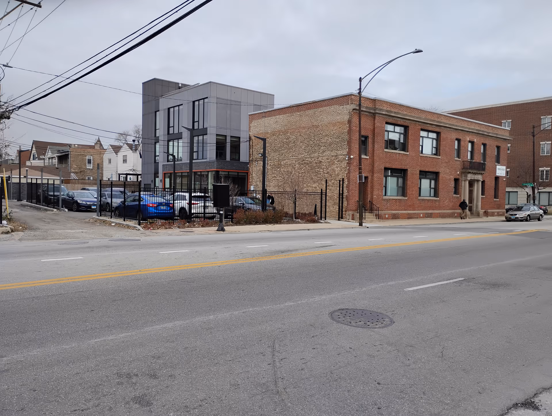 Street view of a brick building with a fenced parking lot and a modern gray building.