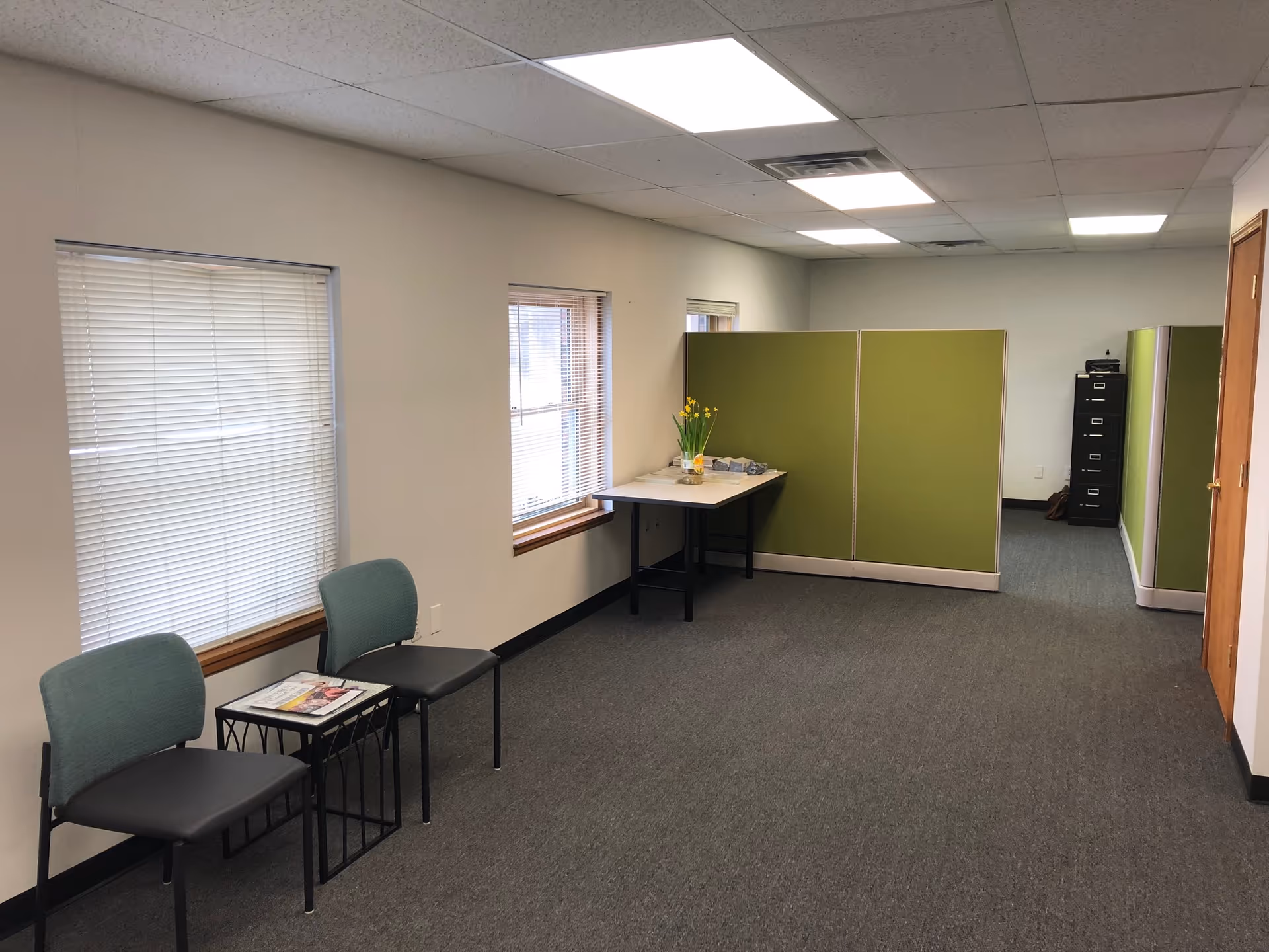 Sparse interior room with green partition, two chairs, a table, and window blinds