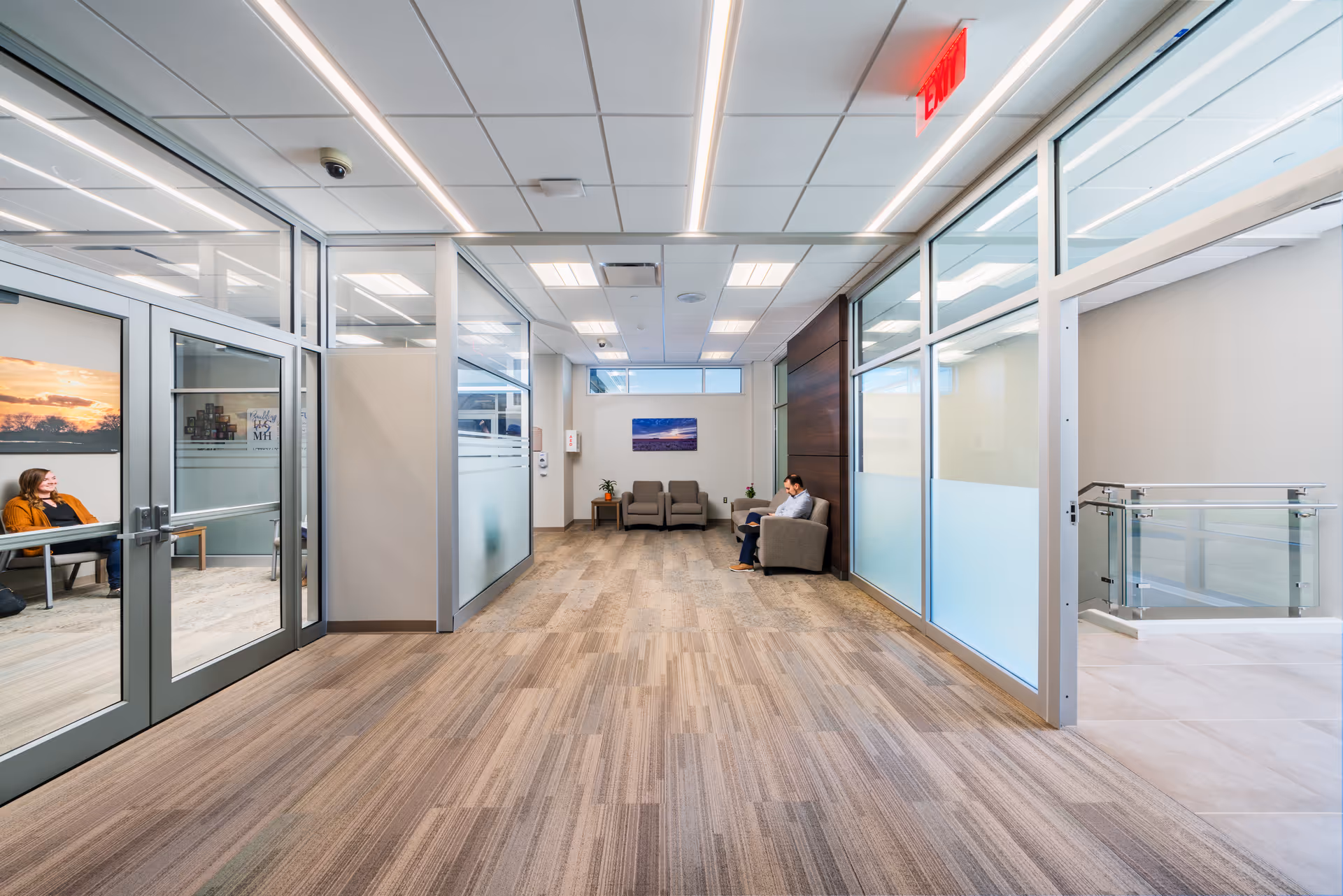 A bright modern hospital hallway with glass-walled offices and a seating area at the end.
