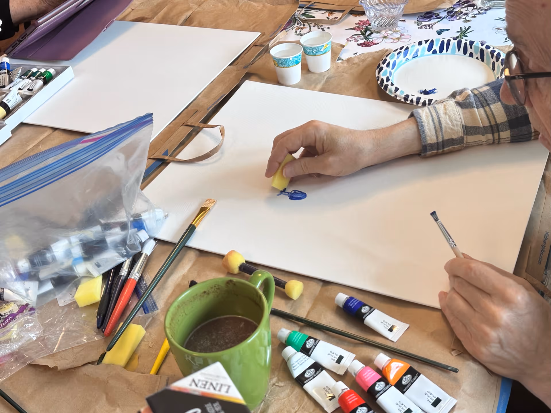 Adult paints on white paper at a cluttered craft table with supplies.