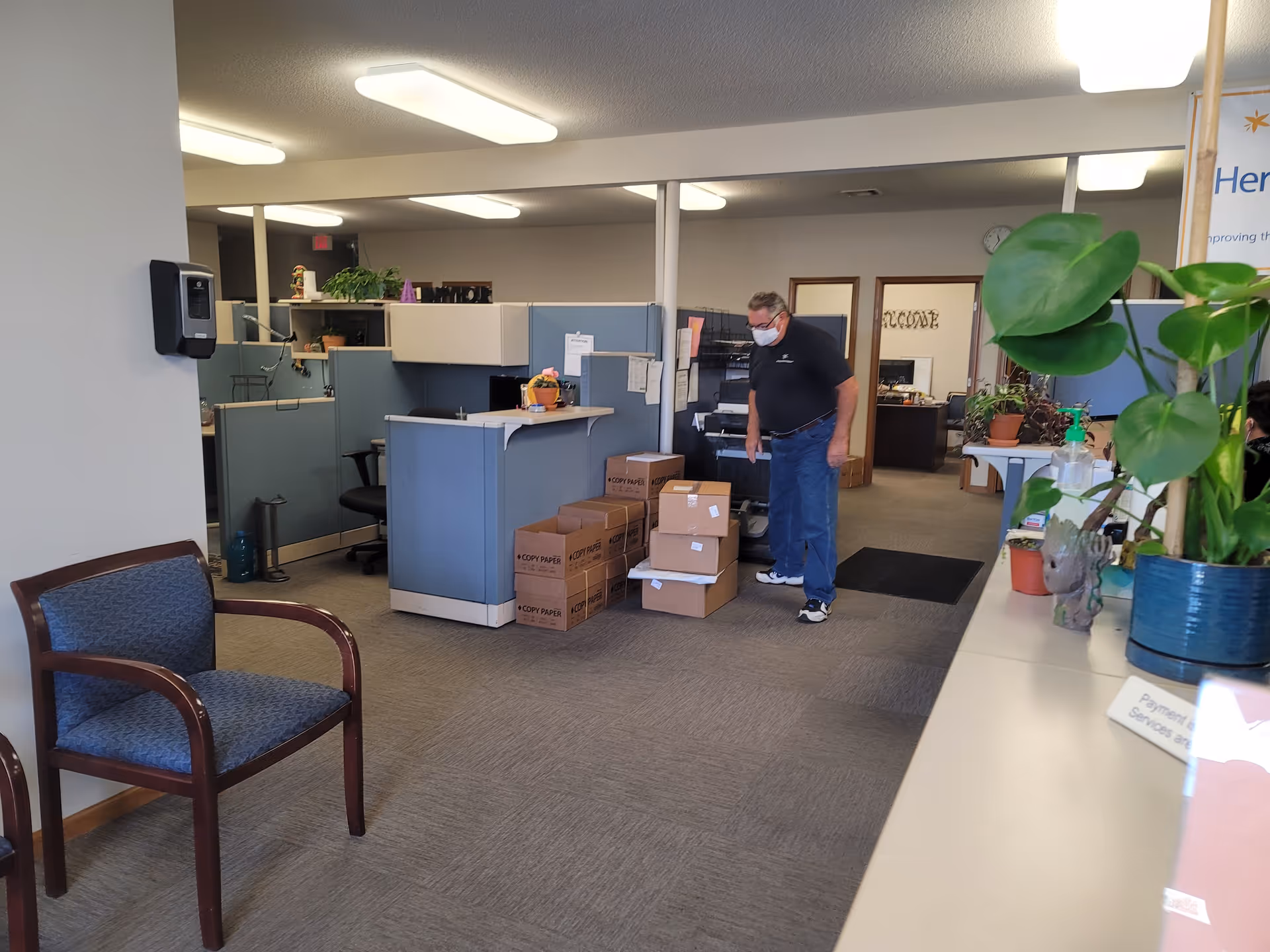 Office reception area with cubicles, boxes, and a masked man inspecting packages.