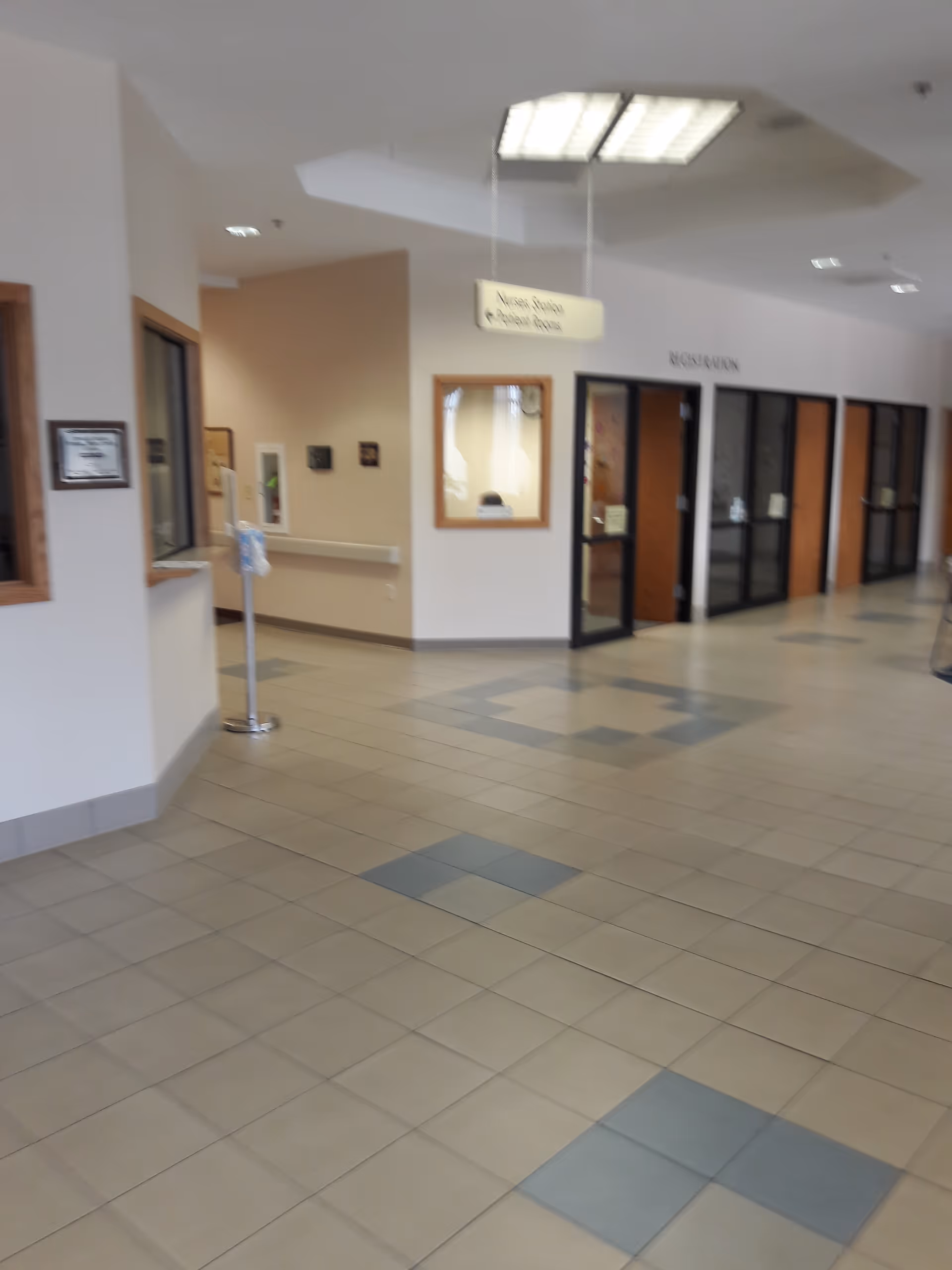 Hospital-like interior hallway with tiled floor, reception windows, and glass doors.