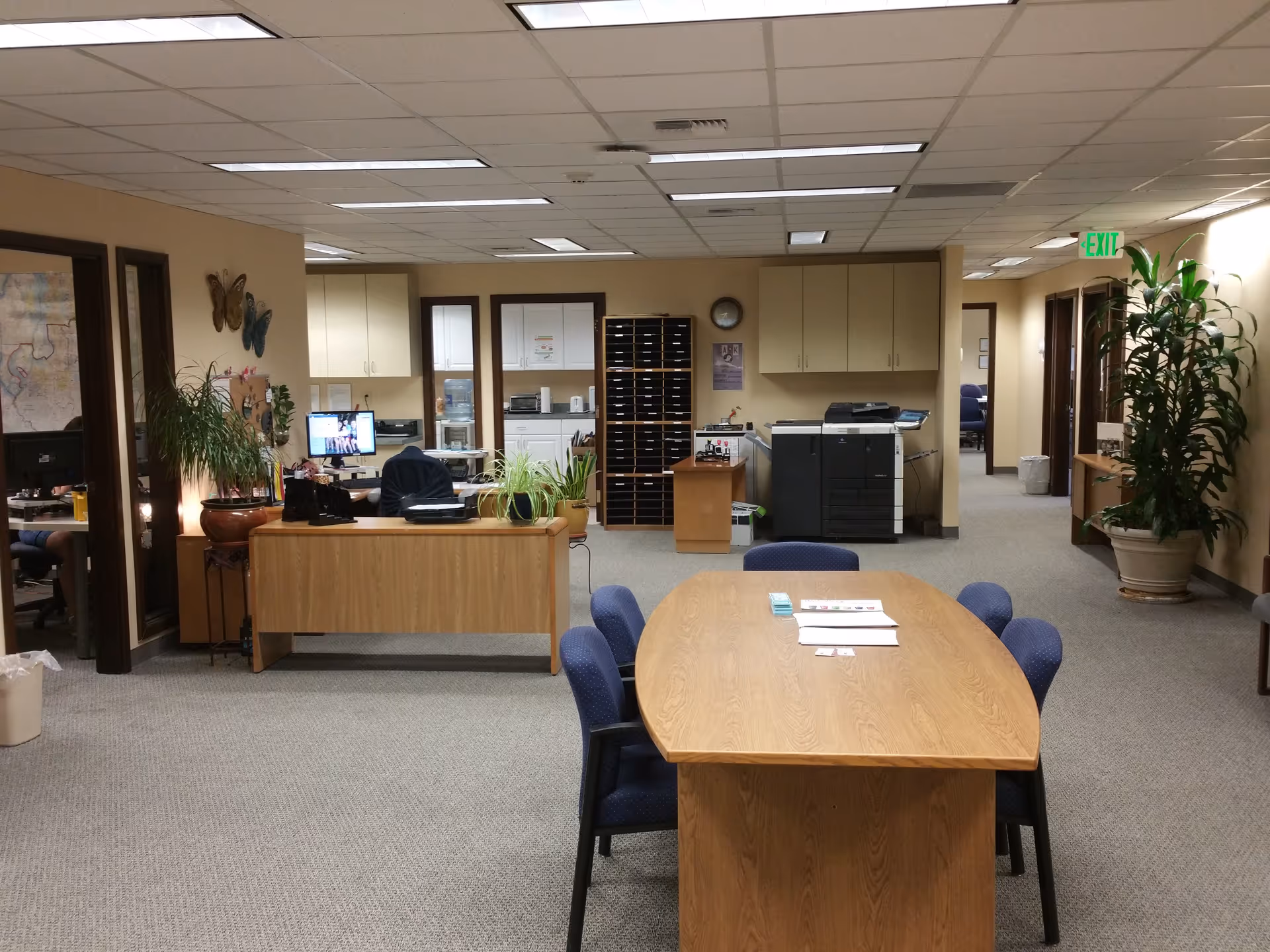 Lobby-style interior with a central wooden table, reception desk, and office equipment.