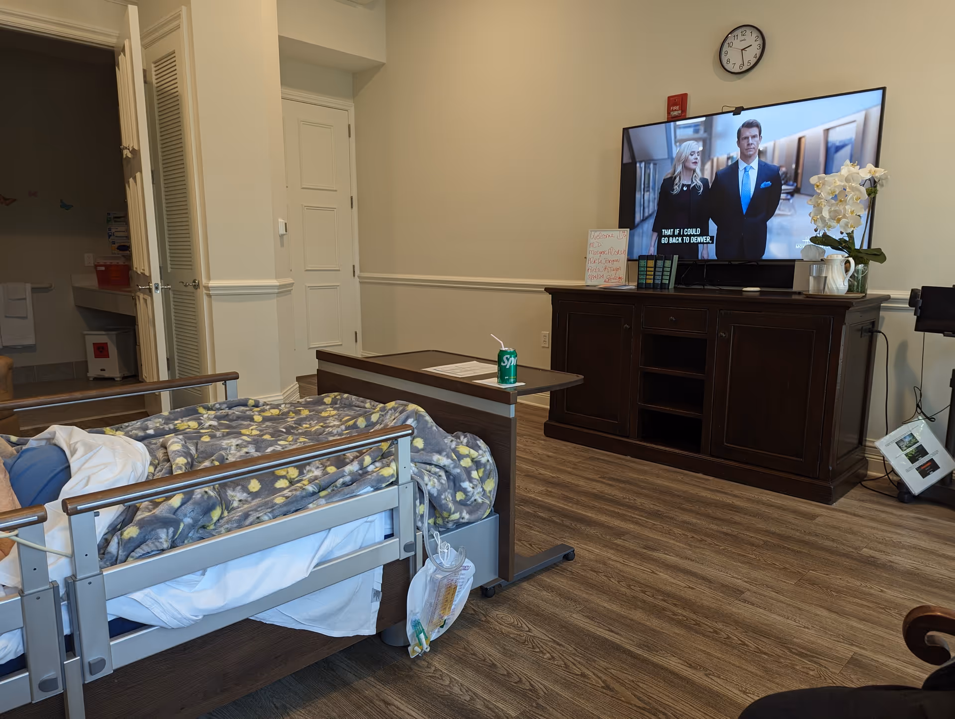 Senior care bedroom with a hospital bed, patterned blanket, and a TV on a dark cabinet.