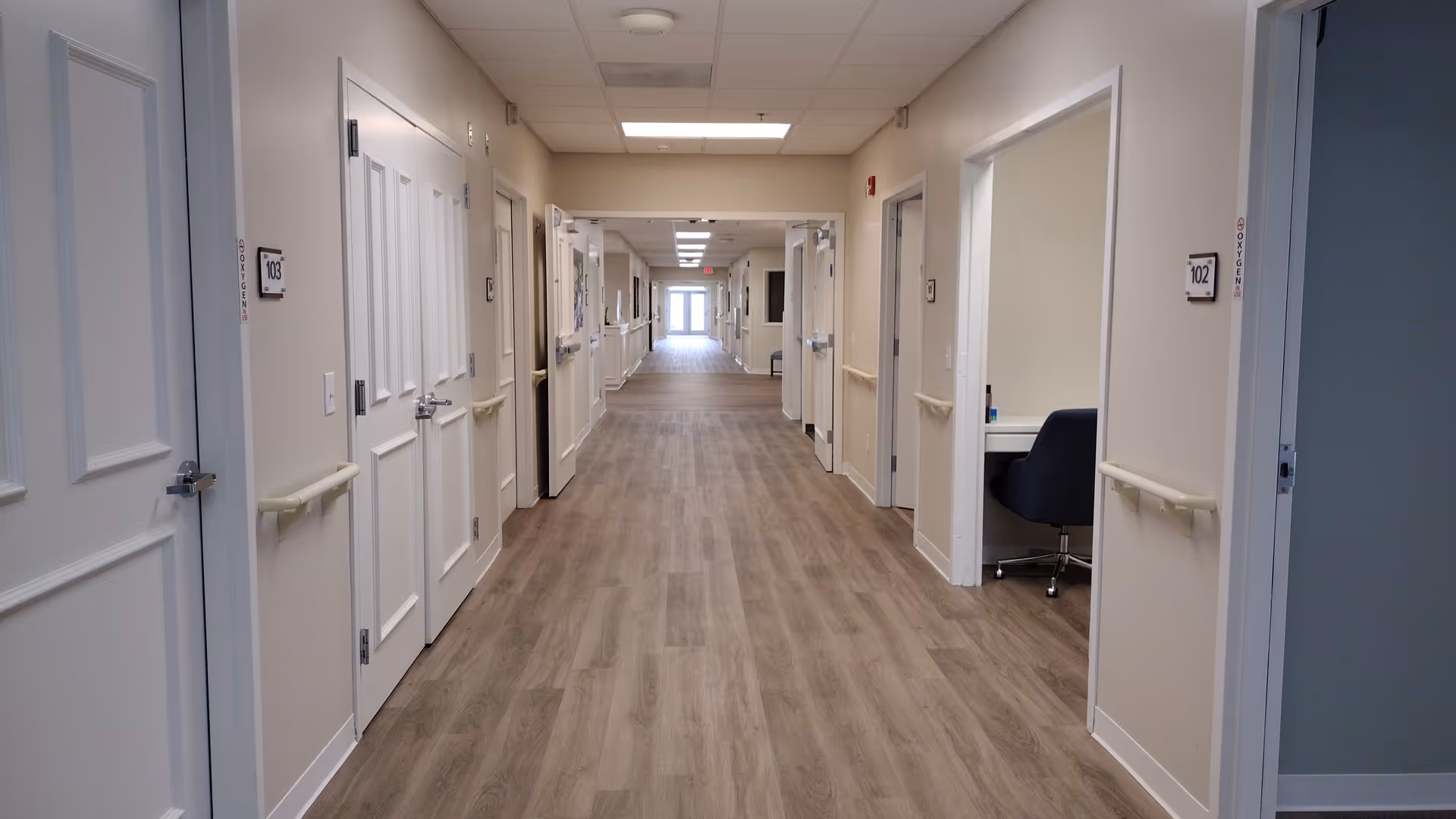Long hallway in a senior care facility with doors on both sides and beige walls.